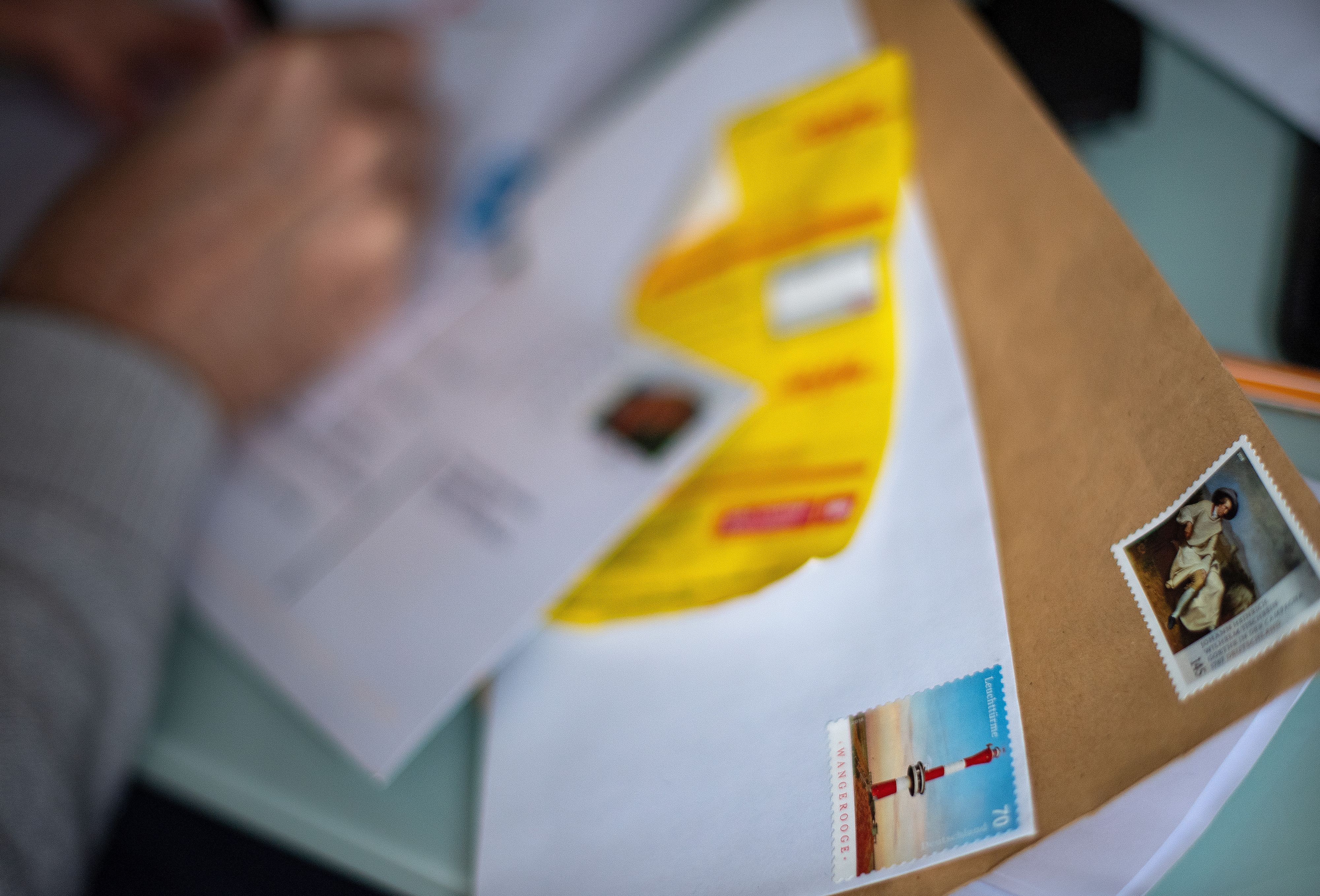Frankierte Briefumschläge liegen auf einem Tisch, während im Hintergrund ein Mann einen Brief adressiert