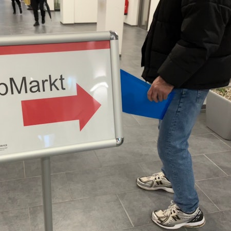 In der Agentur für Arbeit in Köln weist ein Schild auf den Jobmarkt für Geflüchtete hin. Ein Mann mit einer Bewerbungsmappe folgt dem Hinweispfeil.