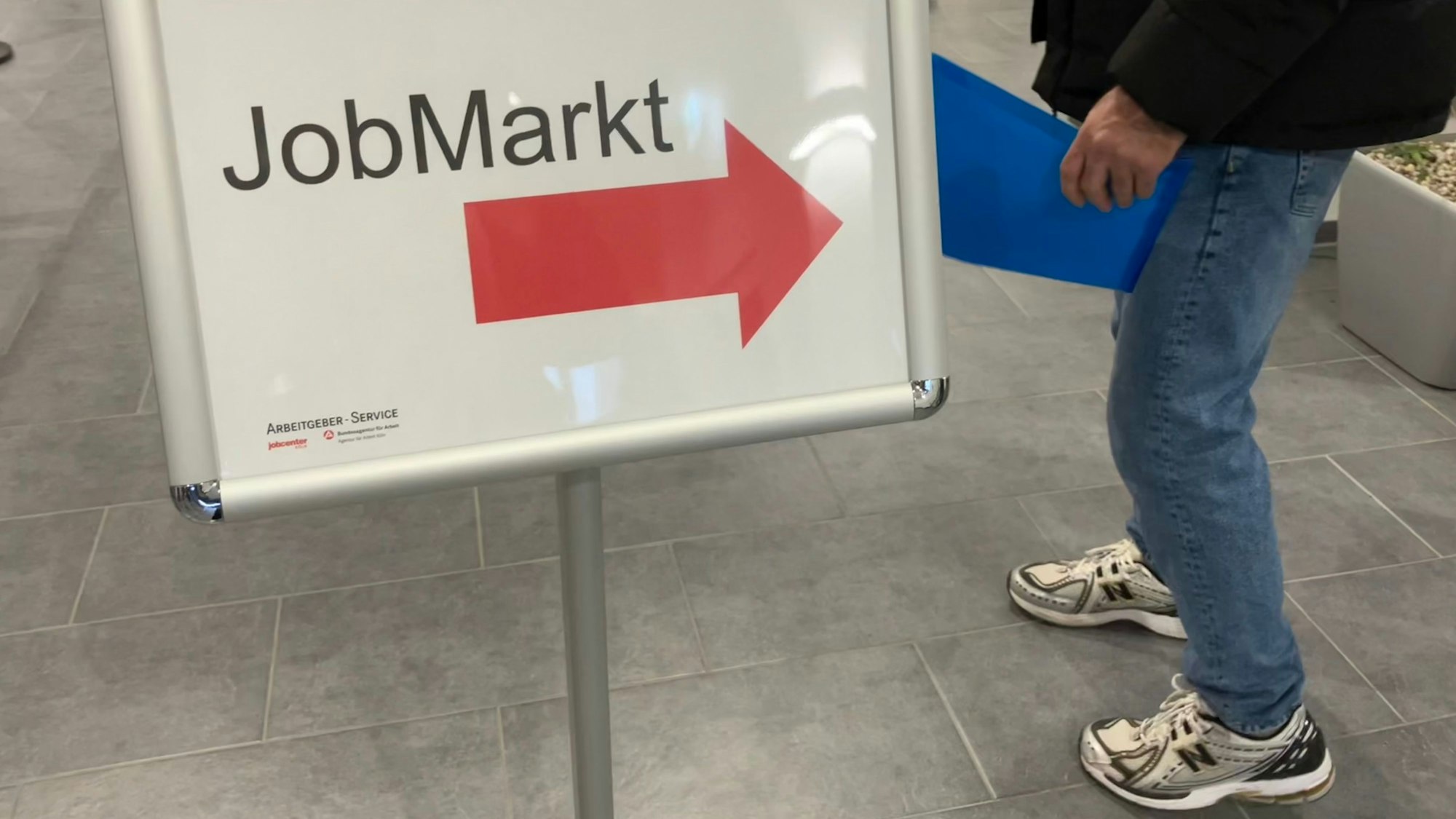 In der Agentur für Arbeit in Köln weist ein Schild auf den Jobmarkt für Geflüchtete hin. Ein Mann mit einer Bewerbungsmappe folgt dem Hinweispfeil.