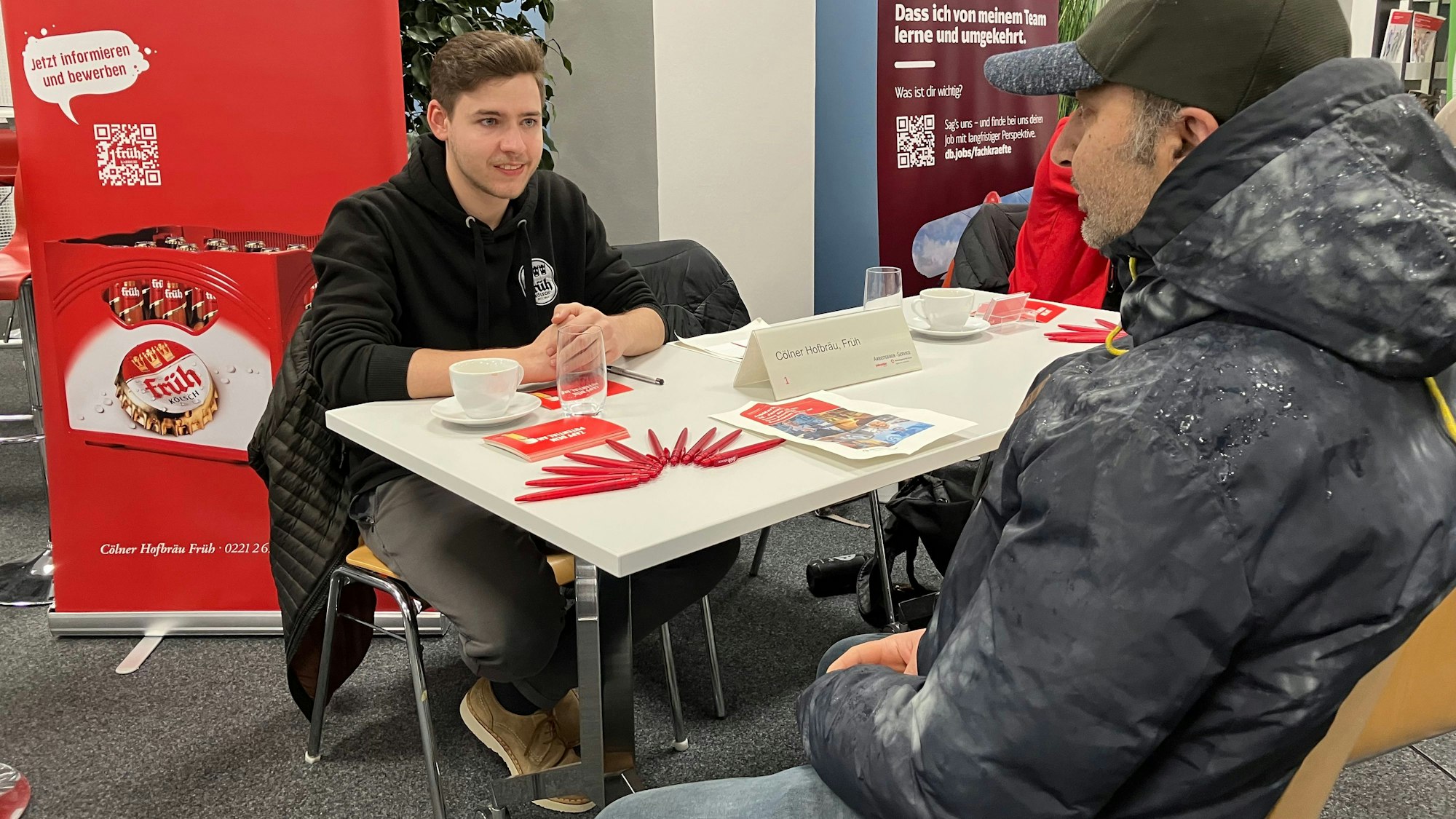 David Pipke, junger Mann in Sweatshirt, sitzt an einem Tisch mit Broschüren, vor einem Plakat von Früh. Ihm gegenüber nimmt Mehmet Cakallikoglu, Mann mittleren Alters mit Cappie, Platz.