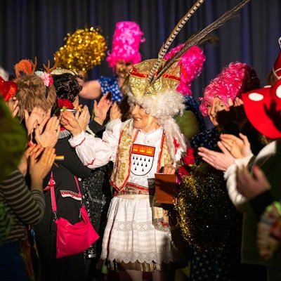 Eine als Prinz Karneval verkleidete Schauspielerin geht durch die Reihen des Publikums.