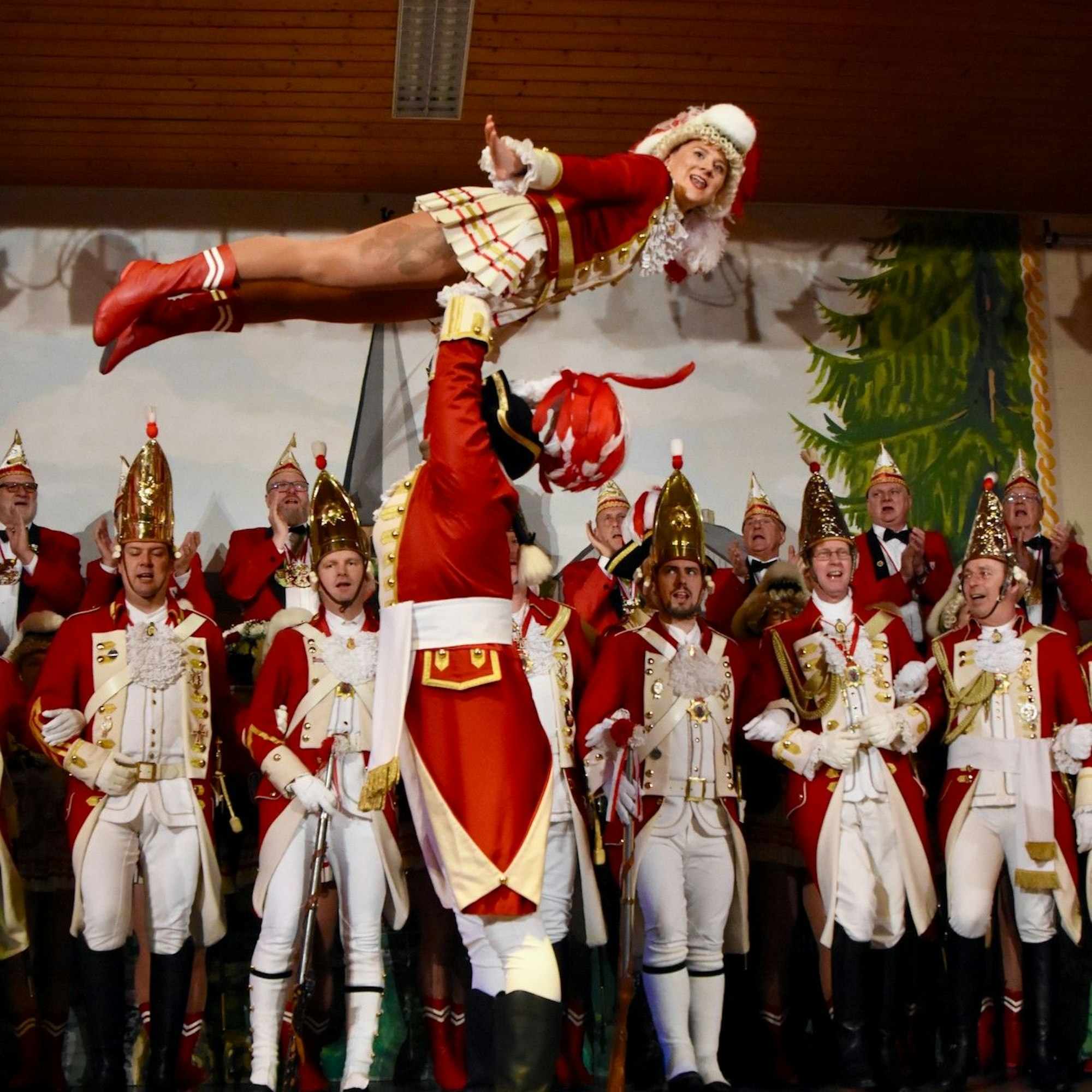 Ein Karnevalstanzpaar mit einer Hebefigur. Im Hintergrund stehen Gardemitglieder in roten und weißen Uniformen.