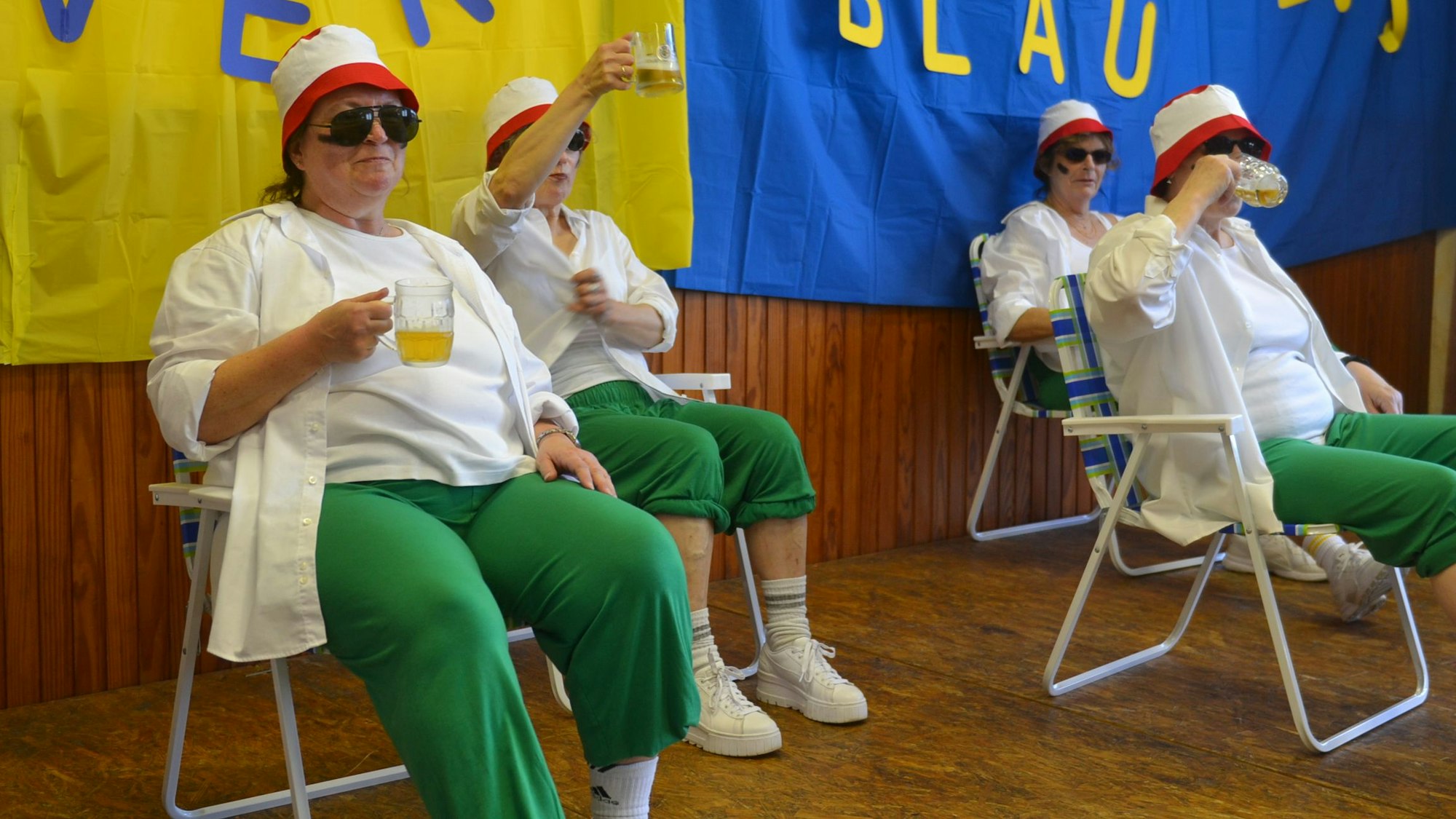 Vier Frauen sitzen mit Sonnenhüten und Bier in Klappstühlen auf einer Bühne.