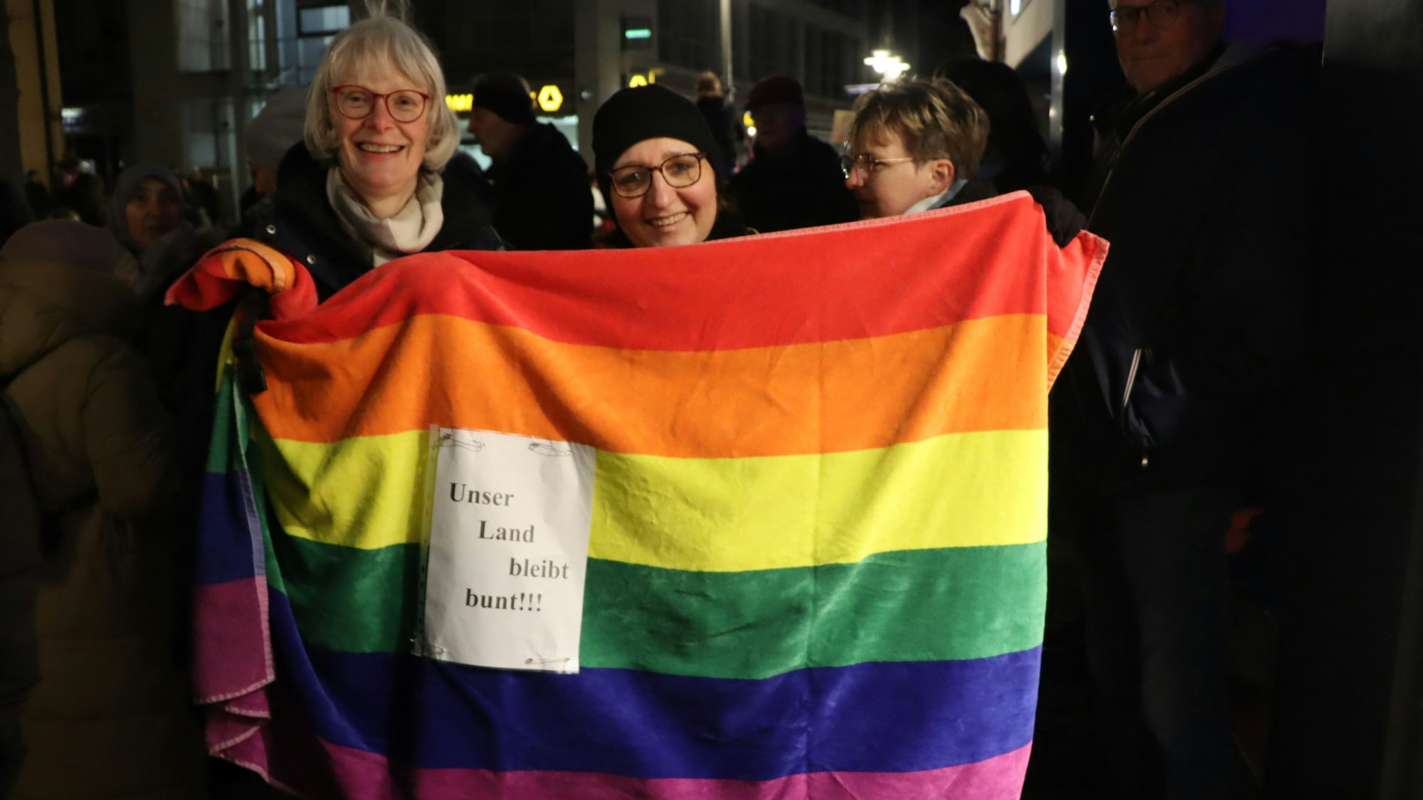 Frauen halten eine Regenbogenfahne hoch, darauf steht „Unser Land bleibt bunt!“.