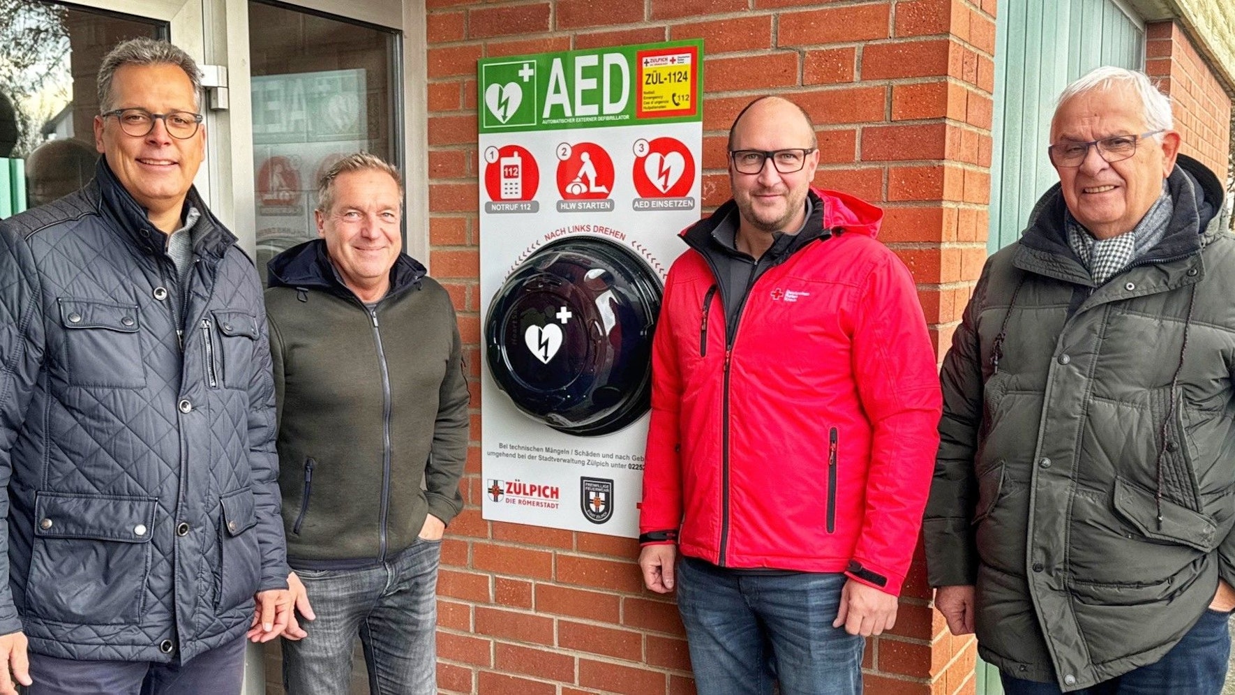 Bürgermeister Ulf Hürtgen steht links im Bild. Er präsentiert mit drei weiteren Männern den neuen Defibrillator an der Dieter-Pritzsche-Halle in Zülpich.