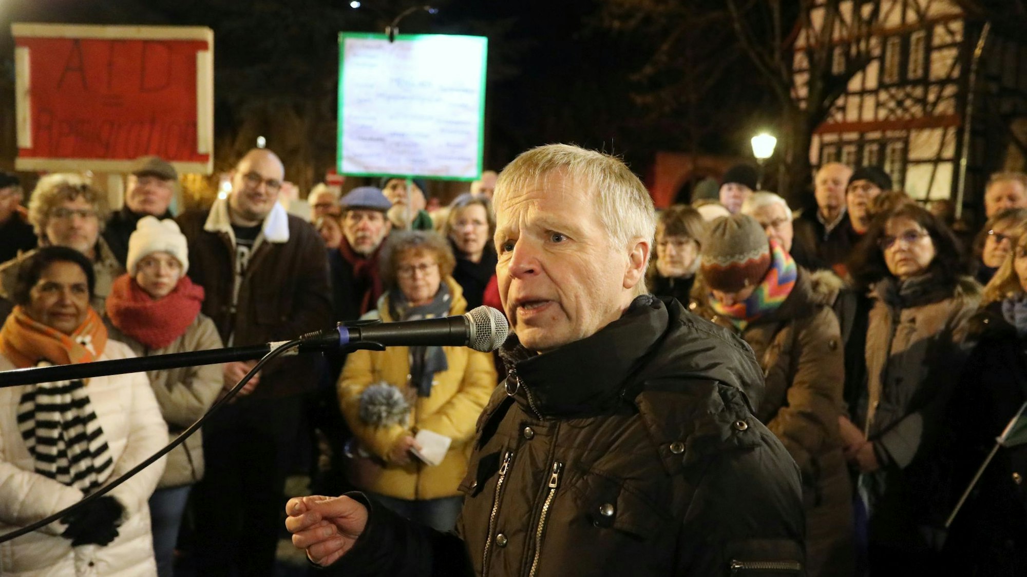 Ein Mann in schwarzer Winterjacke spricht zu Teilnehmern einer Versammlung auf einem öffentlichen Platz.