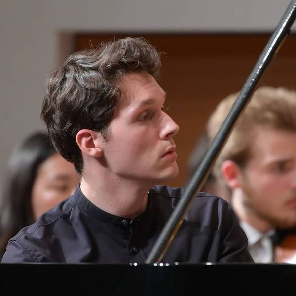 Das Bild zeigt den Pianisten Lukas Sternath 2023 bei einem Konzert des Orchesters der Angelika Prokopp Sommerakademie der Wiener Philharmoniker. Er sitzt am Klavier und schaut auf die rechte Bildseite, sein Gesicht ist im Profil zu sehen. Im Hintergrund zwei Streicher.
