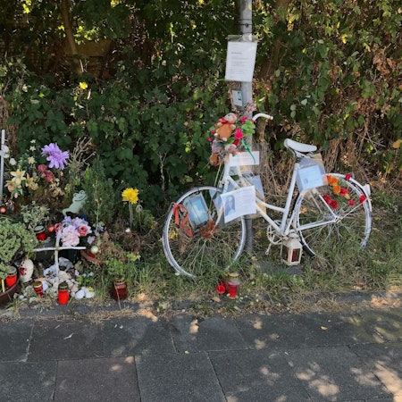 Ein weißes Fahrrad, Blumen und Kerzen stehen am Wegesrand.