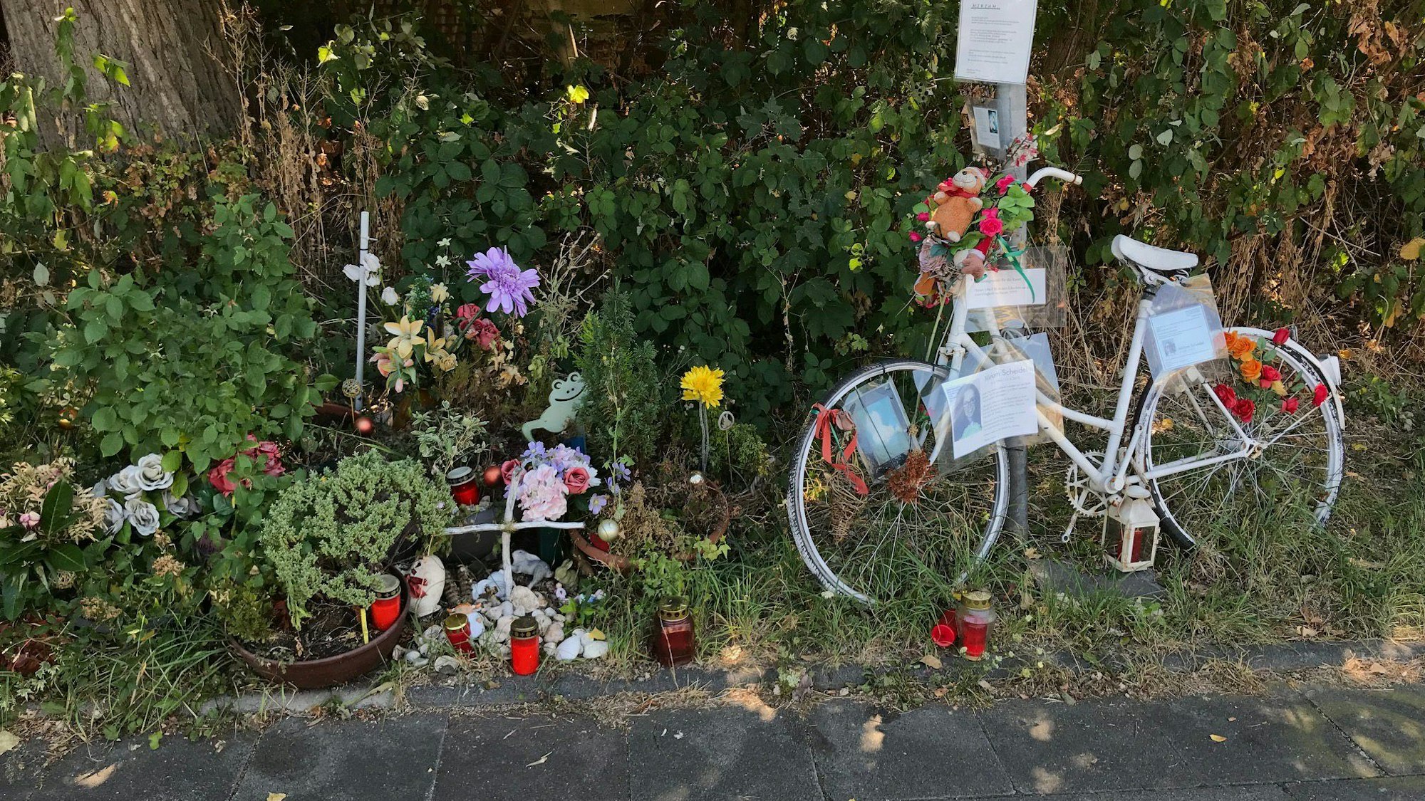 Ein weißes Fahrrad, Blumen und Kerzen stehen am Wegesrand.