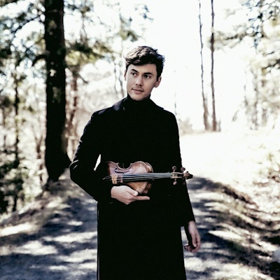 Violinist Benjamin Beilman steht auf einem Weg im Wald. Er hält seine Violine und trägt schwarze Kleidung.