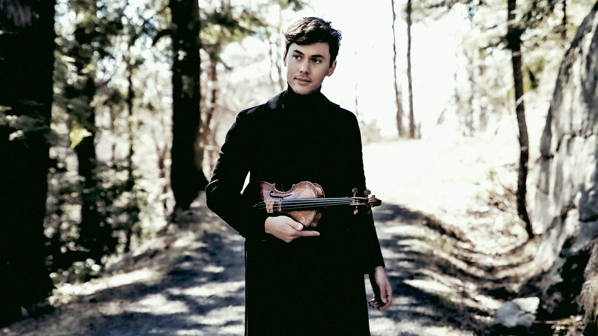 Violinist Benjamin Beilman steht auf einem Weg im Wald. Er hält seine Violine und trägt schwarze Kleidung.
