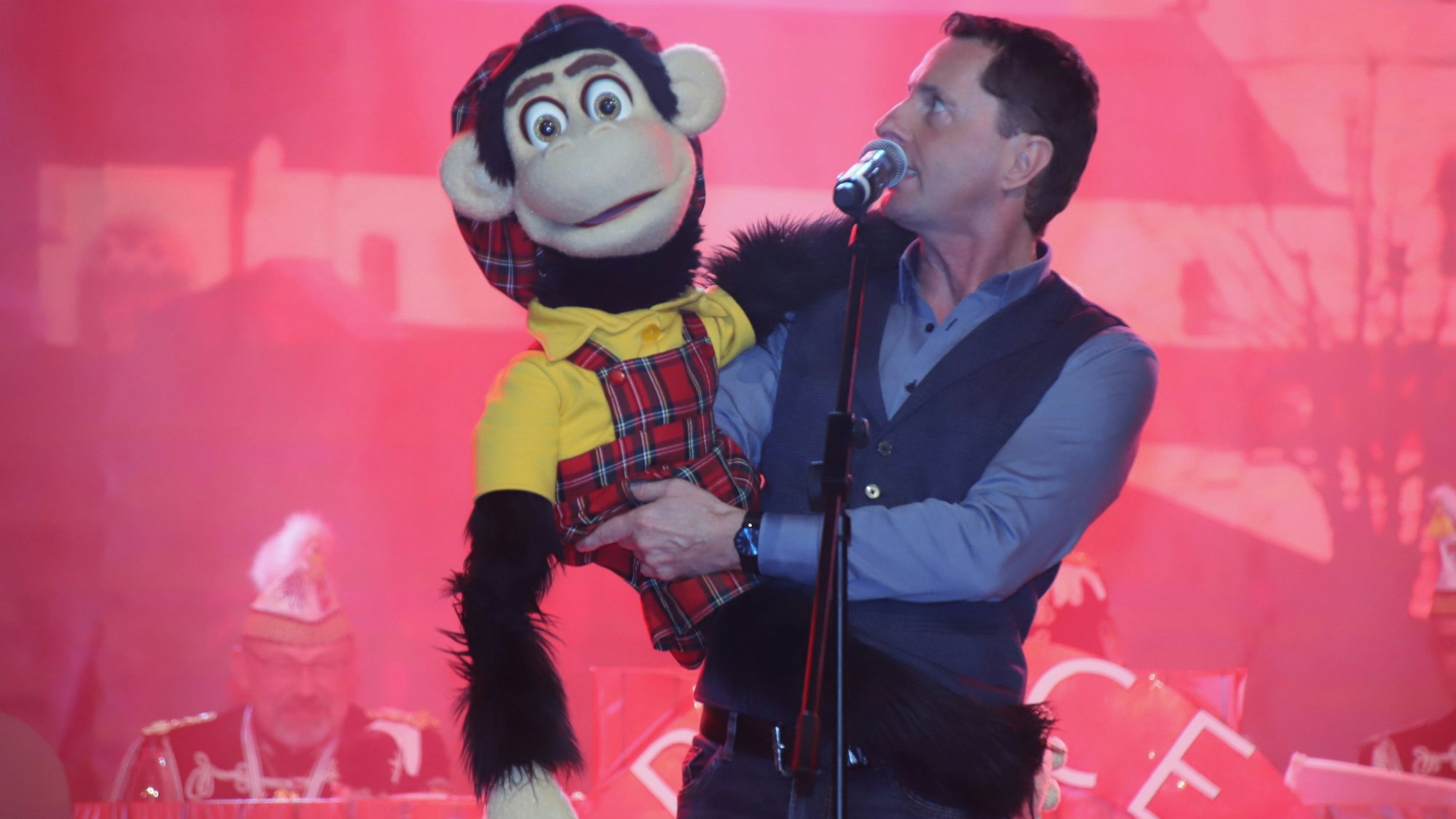 Ein Mann steht an einem Mikrofon, auf einem Arm hält er einen großen Stoffaffen in karierter Latzhose, den er anschaut.