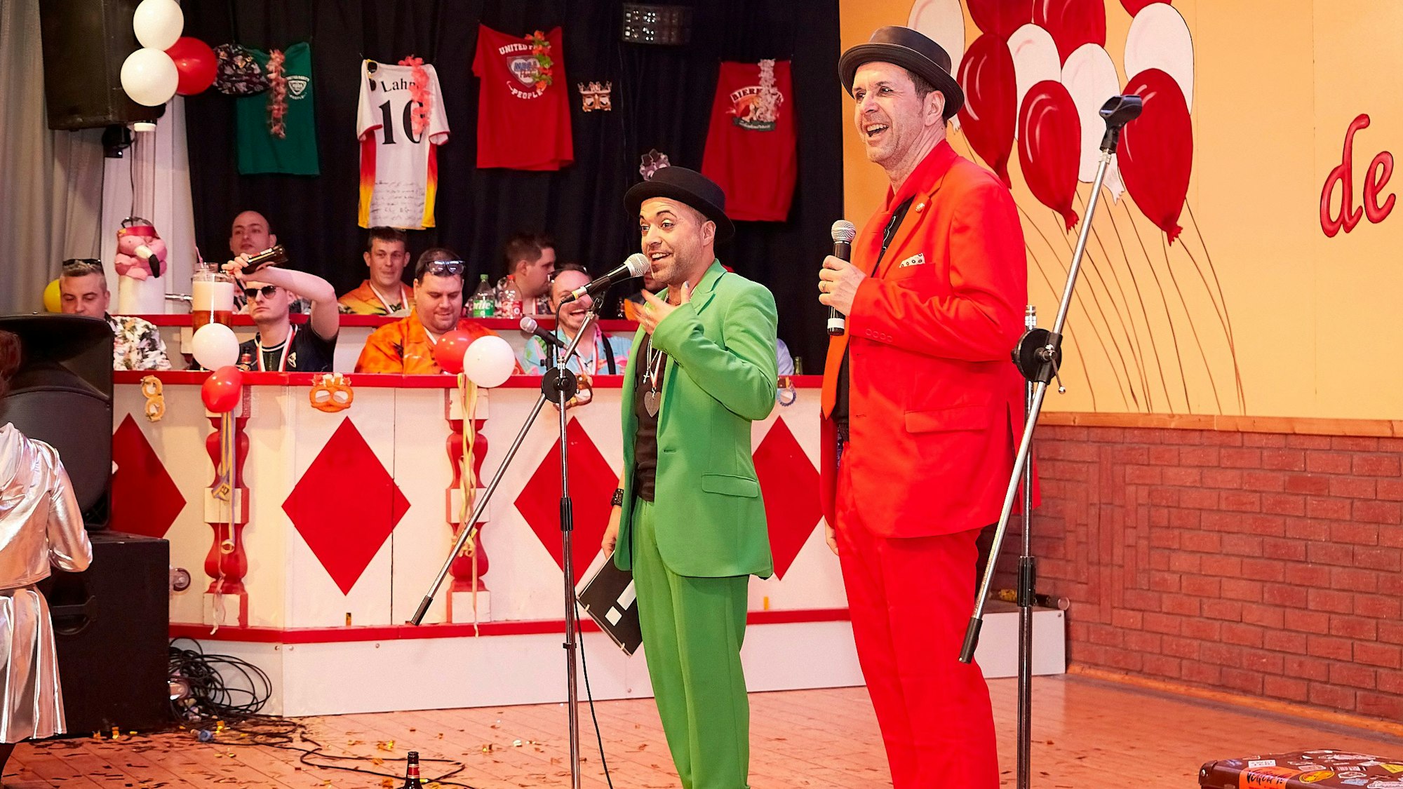 Zwei Redner, einer in rotem Anzug, einer in grünem Anzug, stehen auf einer Karnevalsbühne vor Mikrofonen.