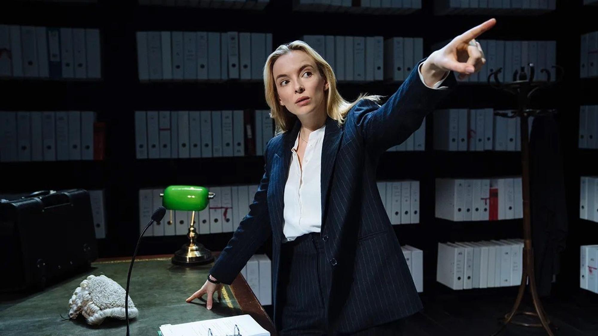Die englische Schauspielerin Jodie Comer in „Prima facie“, ein Ein-Personen-Stück am Londoner West End und am New Yorker Broadway. Sie steht an einem Schreibtisch vor Aktenregalen, gestikuliert mit ausgestrecktem Arm und Zeigefinger.
