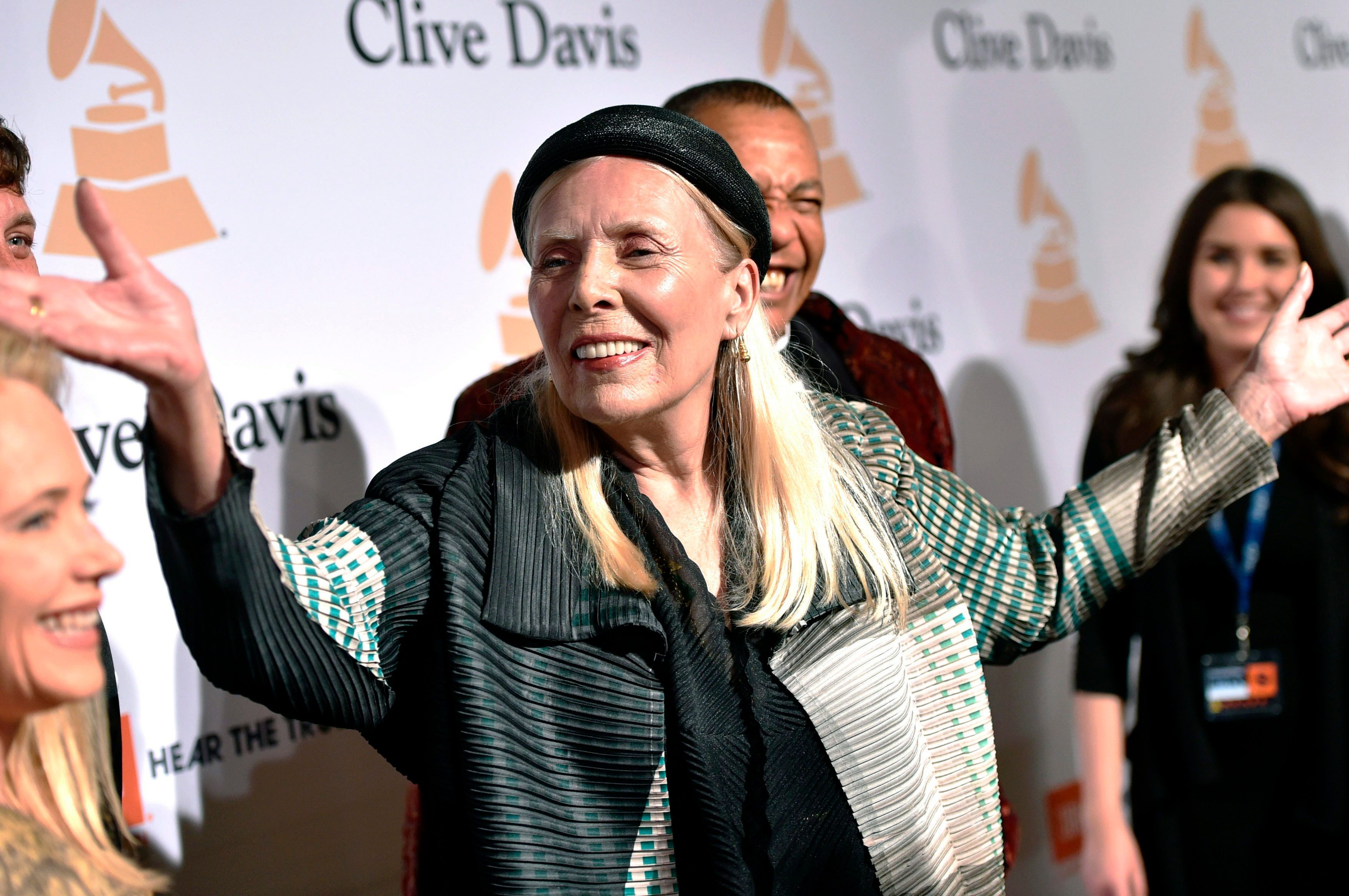 In dem Bild ist die zu dem Zeitpunkt 72-jährige Joni Mitchell auf dem roten Teppich zu sehen. Sie hat die Arme ausgebreitet und lächelt.