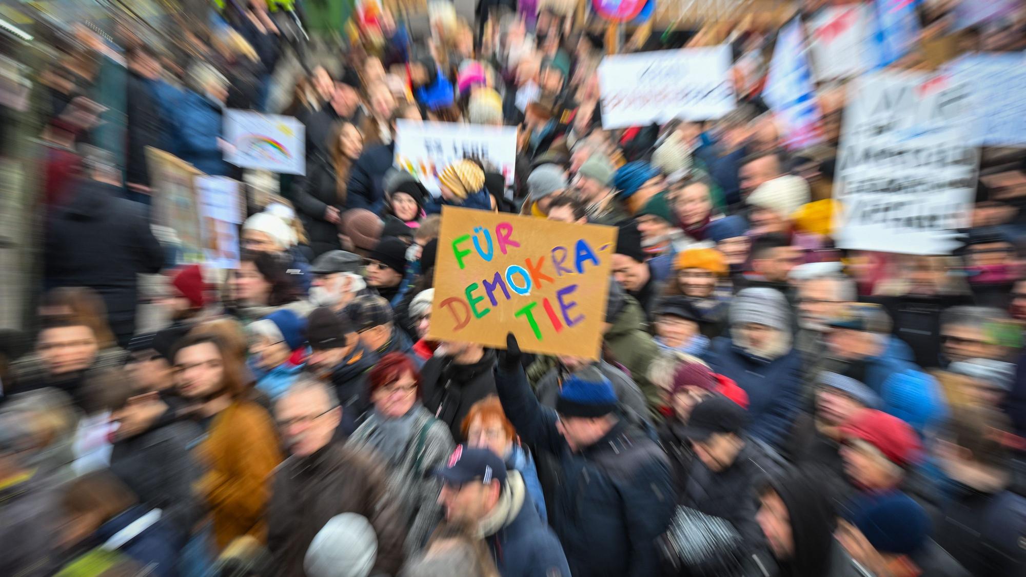 Das Bild zeigt Teilnehmer einer Demonstration, die Plakate hochhalten. Während alle Personen und Schilder verzerrt dargestellt sind, ist nur ein Plakat in der Mitte des Fotos scharf abgebildet. Auf ihm ist zu lesen „Für Demokratie“.