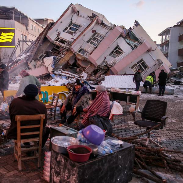 Im türkischen Ort Golbasi in der Provinz Adiyaman sitzen im Februar 2023 Menschen auf Stühlen auf der Straße. Im Hintergrund ist ein eingestürztes Gebäude zu sehen.