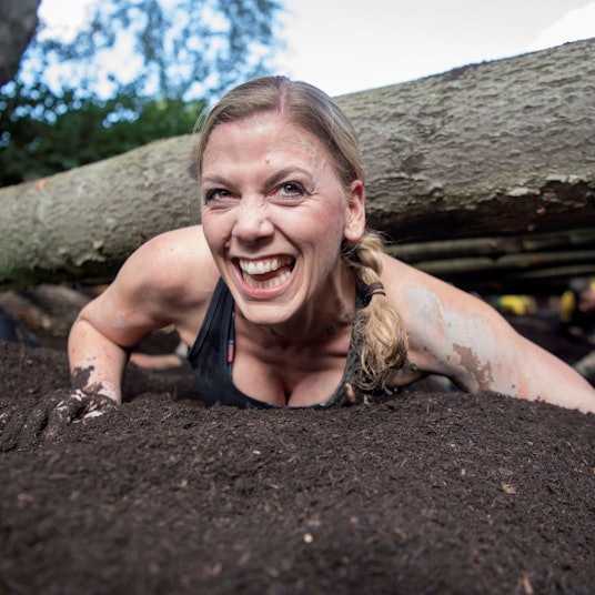 Das Bild zeigt eine Teilnehmerin, die unter Baumstämmen durch robbt und dabei in die Kamera lächelt.