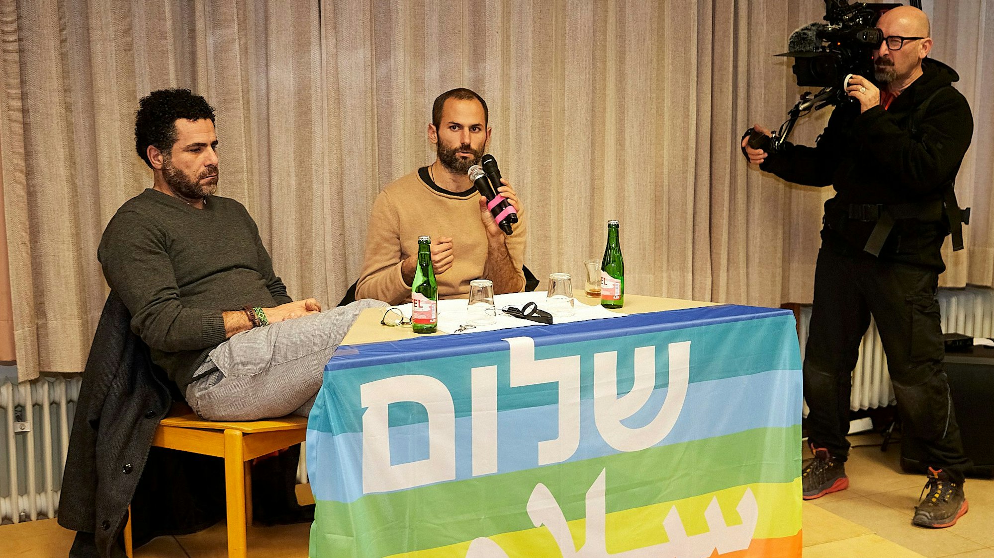 Der israelische Soldat Rotem Levin (Mitte) und der palästinensische Widerstandskämpfer Osama Iliwat sitzen gemeinsam an einem Tisch und werden dabei von einem Kameramann gefilmt.