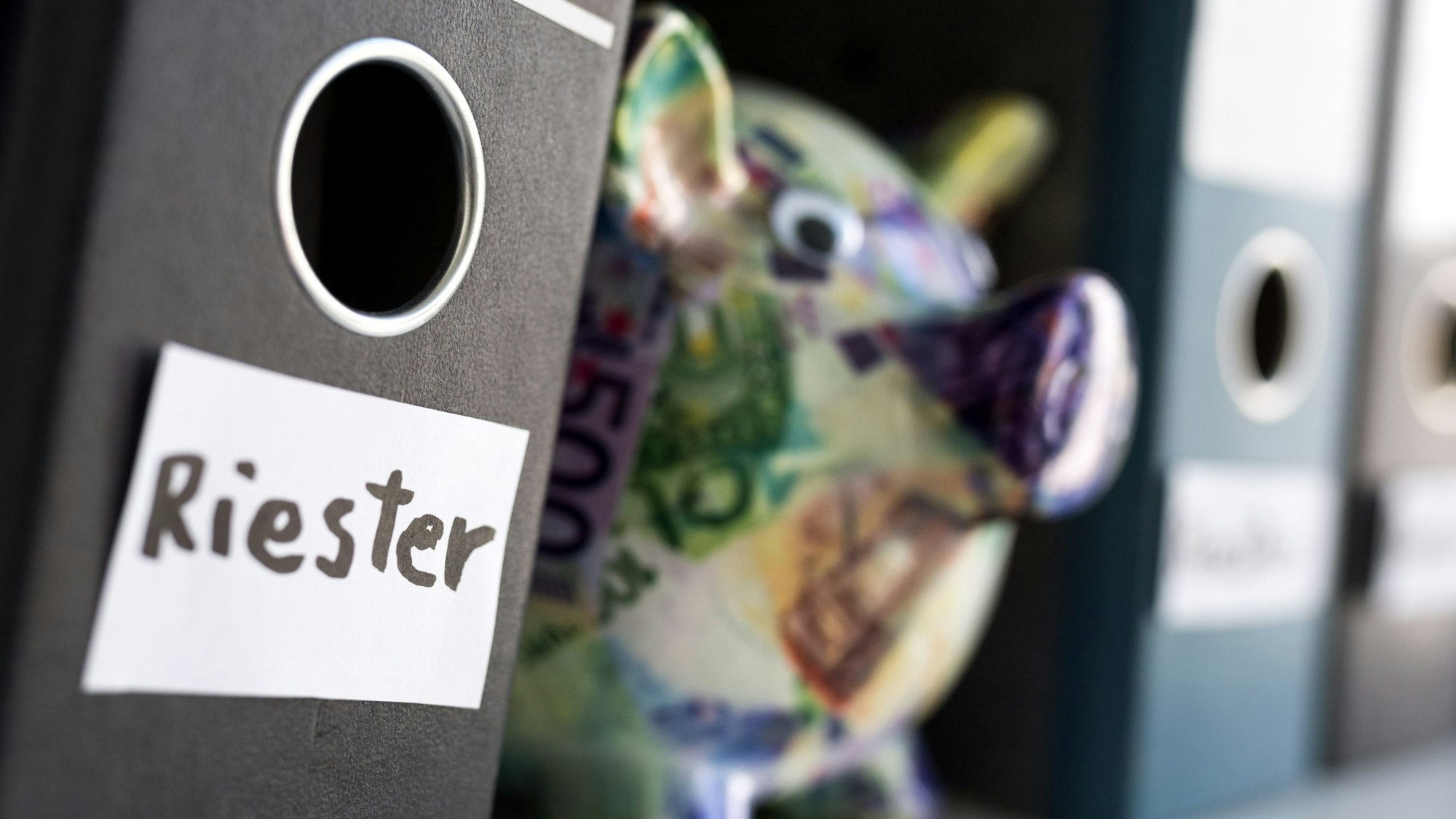 Ein Ordner mit der Aufschrift „Riester“ steht neben anderen Ordnern und einem Sparschwein in einem Regal.