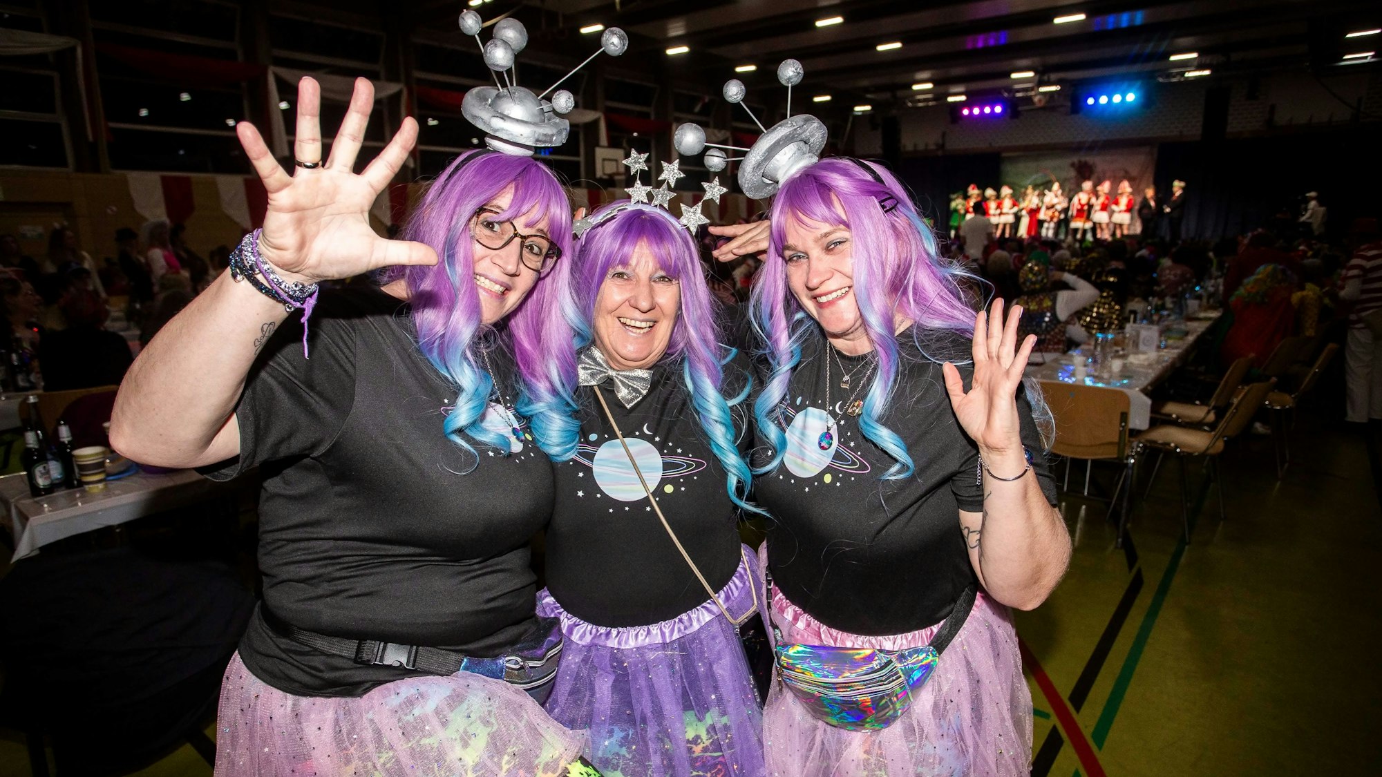 Drei Frauen mit lila Perücken winken in die Kamera.