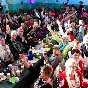 Bunt kostümierte Jecke sitzen in der Dorfhalle Mahlberg eng beisammen an Tischen. Viele lachen und recken zum Alaaf-Ruf die Arme in die Höhe.