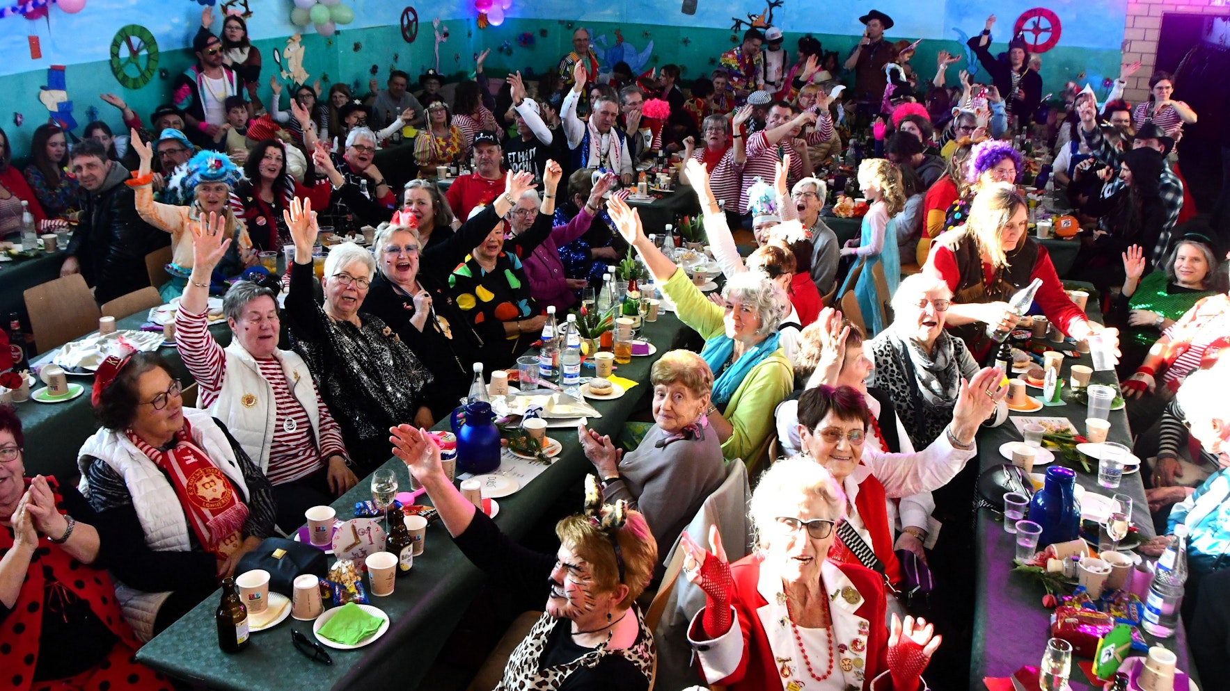 Bunt kostümierte Jecke sitzen in der Dorfhalle Mahlberg eng beisammen an Tischen. Viele lachen und recken zum Alaaf-Ruf die Arme in die Höhe.