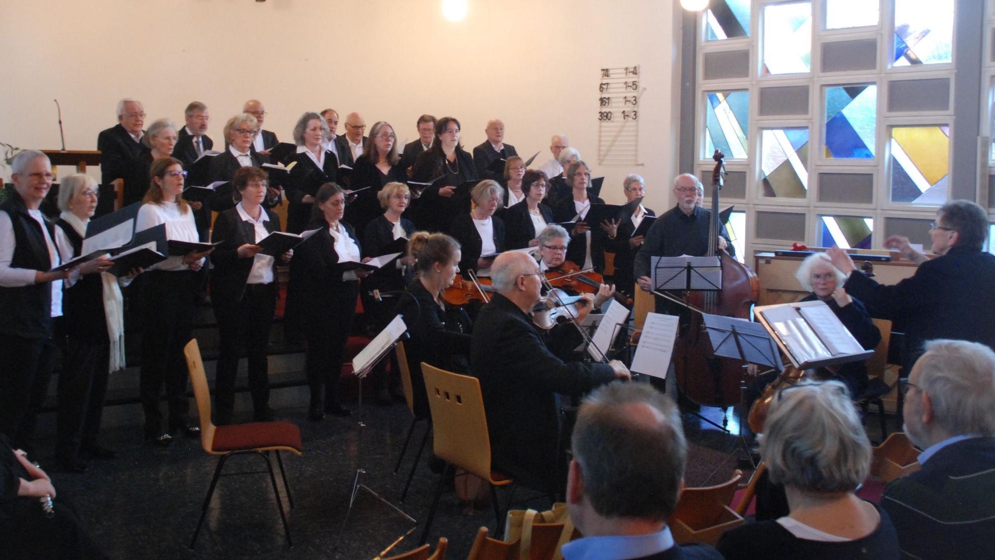 Die Chormitglieder stehen in drei Reihen auf Treppenstufen, vor ihnen ist das Orchester platziert, rechts im Bild die Dirigentin.