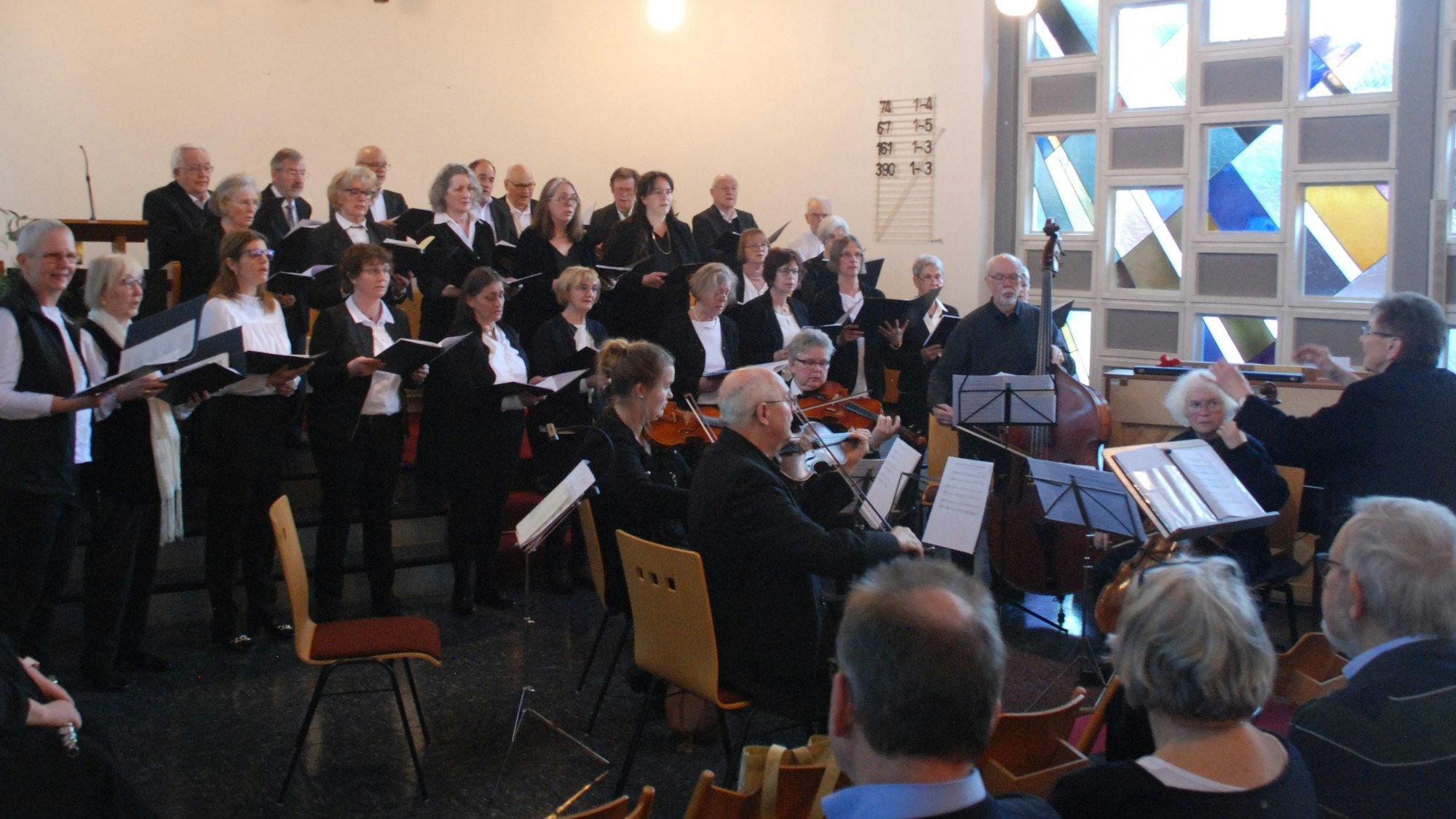 Die Chormitglieder stehen in drei Reihen auf Treppenstufen, vor ihnen ist das Orchester platziert, rechts im Bild die Dirigentin.