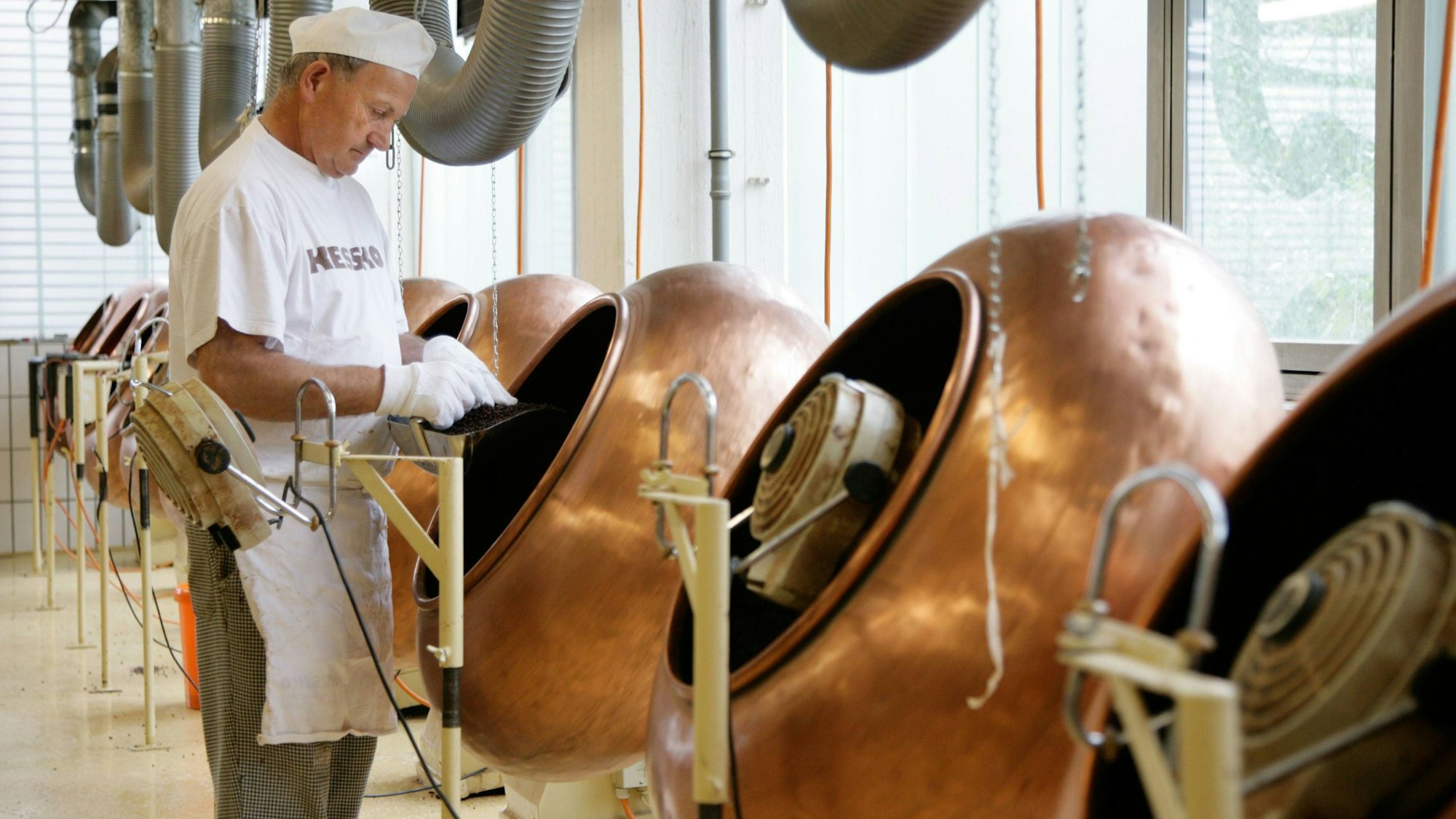 Ein Mann arbeitet an Maschinen, die zur Schokoladenherstellung dienen. Er steht links im Bild und trägt weiße Arbeitskleidung. Die Maschinen sind bronzefarben.