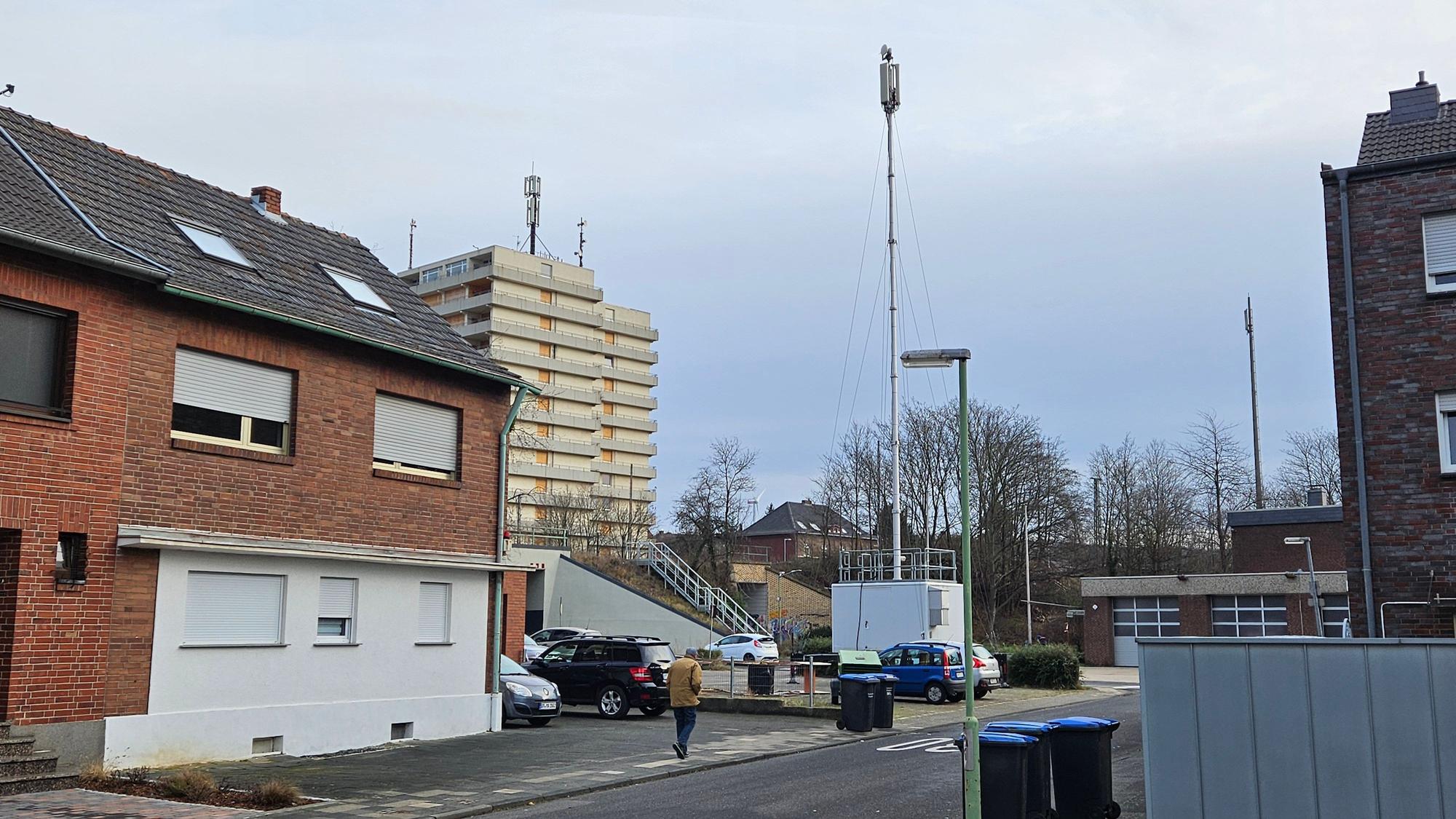 Ein Funkmast steht auf einem Parkplatz zwischen Gebäuden, im Hintergrund ist ein Hochhaus zu sehen.