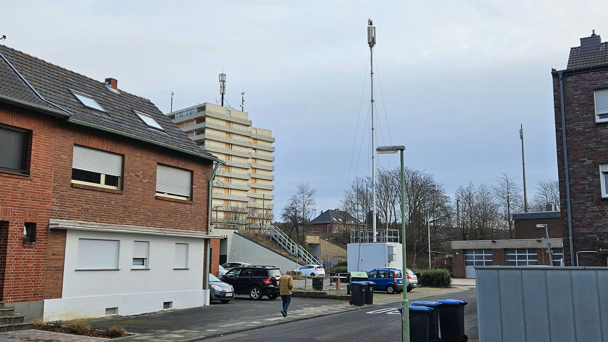 Ein Funkmast steht auf einem Parkplatz zwischen Gebäuden, im Hintergrund ist ein Hochhaus zu sehen.