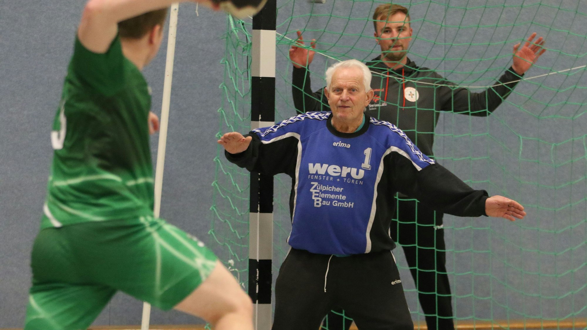 Eckart Wudarzak hat beim Warmmachen den Ball im Blick. Ein Mitspieler setzt zum Sprung an, hinter dem Tor redet ein Mitspieler mit ihm.