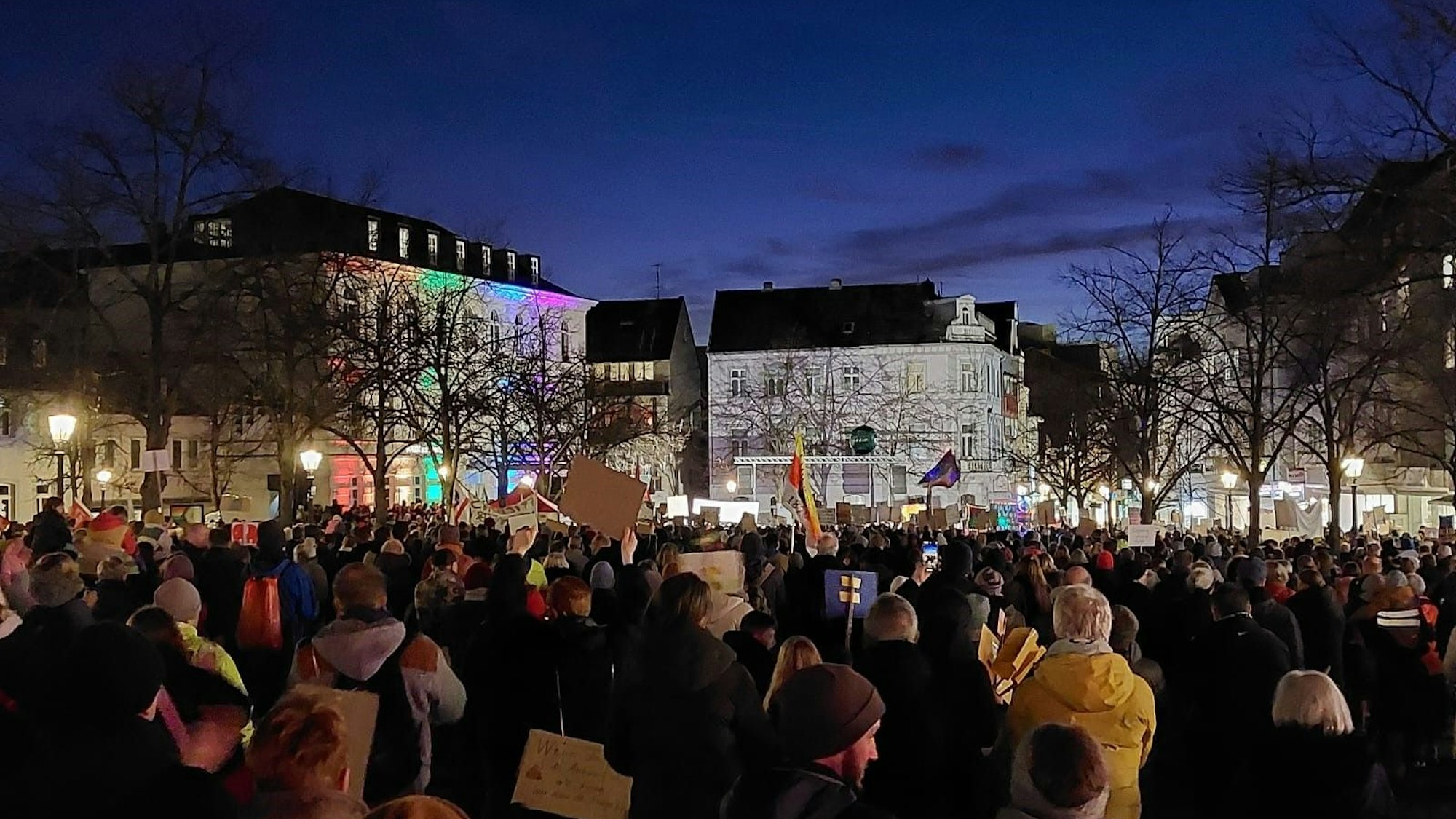 Teilnehmer einer Kundgebung auf einem Marktplatz. Ein weißes Gebäude ist in Regenbogenfarben angestrahlt.