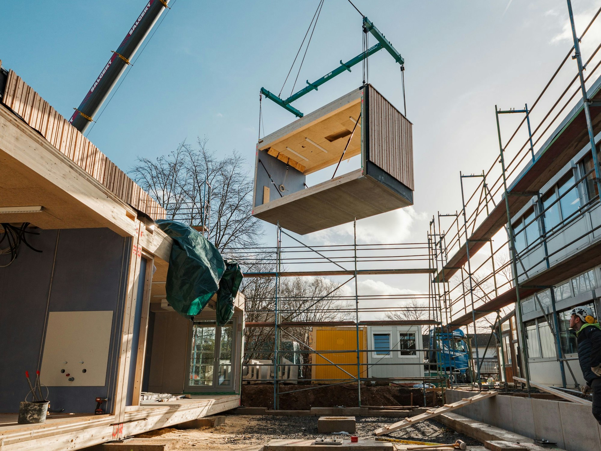 An einem Kran hängt ein Holzbaustein, der Teil eines Schulgebäudes ist.