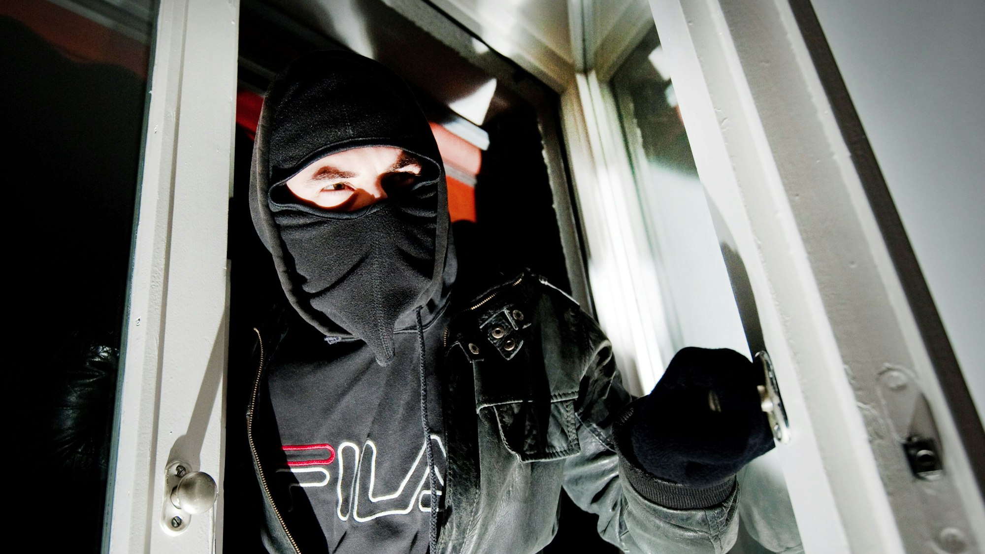 Ein vermummter Einbrecher betritt über eine Terrassentür eine Wohnung.