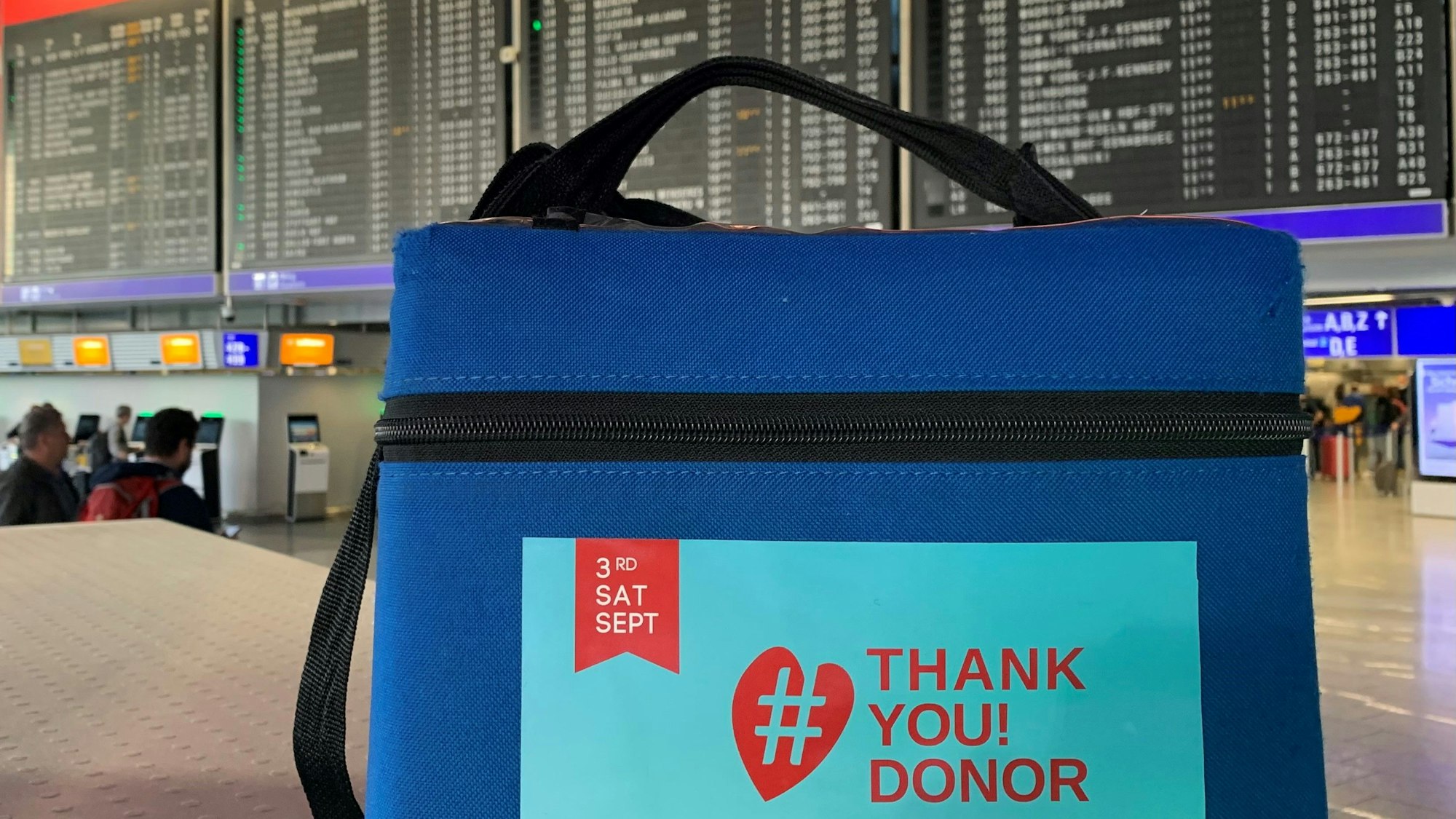 Eine blaue Stammzellentransportbox am Frankfurter Flughafen. Im Hintergrund ist die Fluganzeigetafel zu sehen.