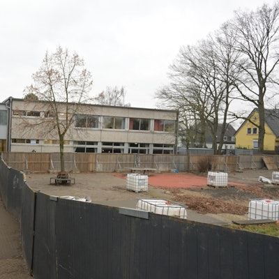 Zwischen dem Schulgebäude und dem Bauzaun sieht man den Gang. Hinter dem Zaun sieht man, den früheren Schulhof.