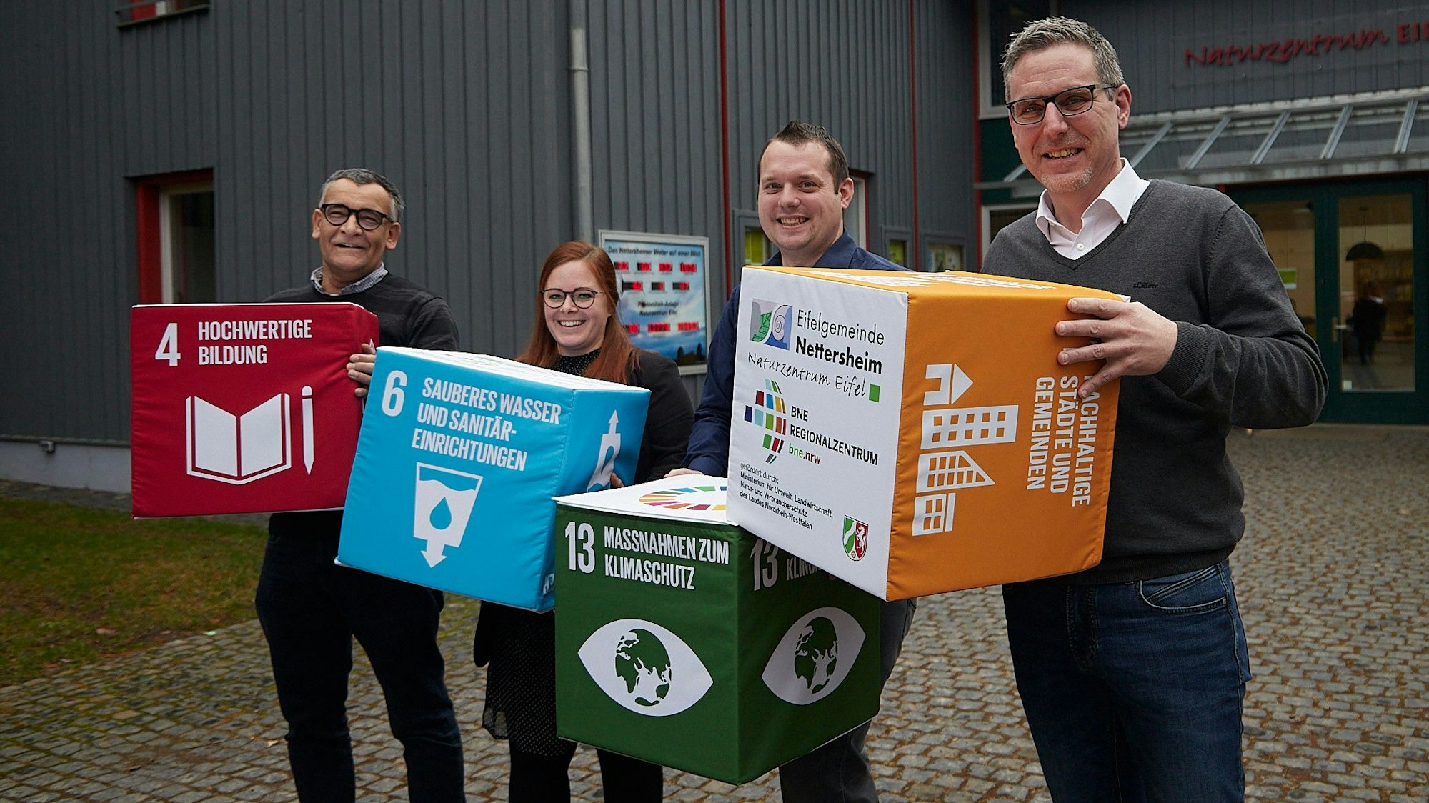Drei Männer und eine Frau stehen vor dem Naturzentrum Nettersheim. Sie halten Würfel in der Hand, auf denen Nachhaltigkeitsziele der Vereinten Nationen stehen.