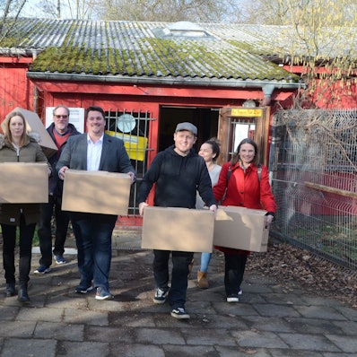 Männer und Frauen vor einem flachen roten Holzbau; sie tragen Umzugskisten.