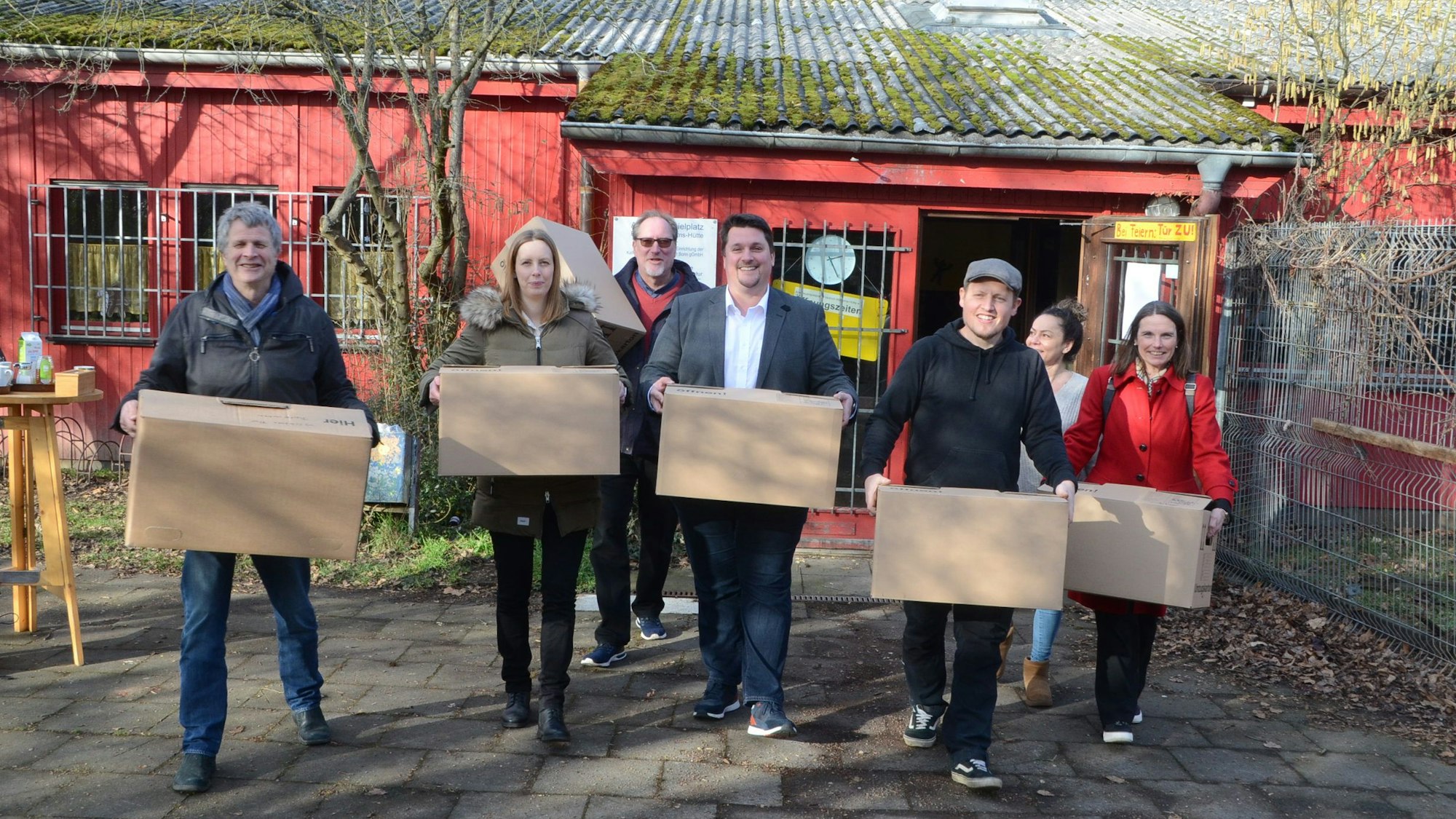 Männer und Frauen vor einem flachen roten Holzbau; sie tragen Umzugskisten.