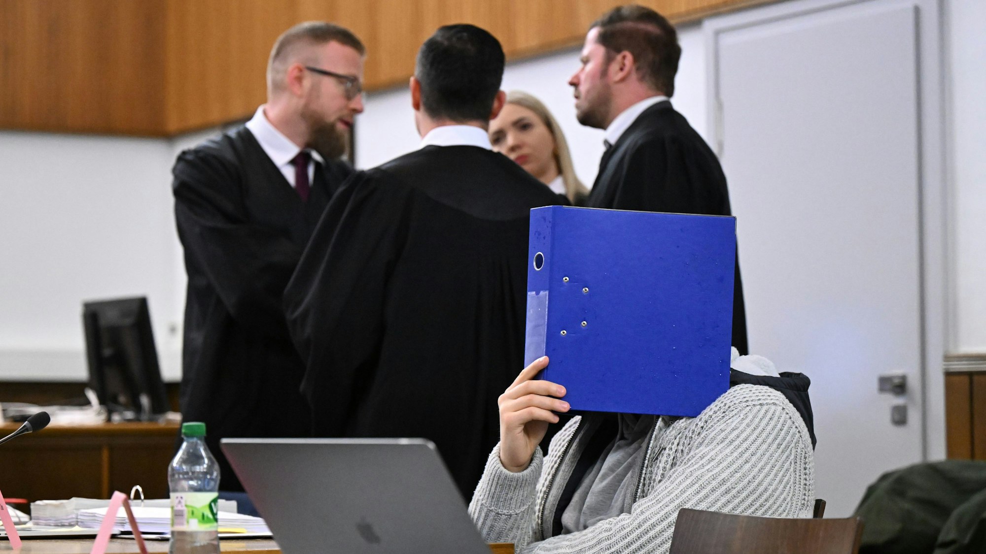 01.02.2024, Baden-Württemberg, Mosbach: Der Mitangeklagte (vorne) sitzt vor Beginn seines Prozesses in einem Gerichtssaal im Landgericht Mosbach, mit einem Aktenordner vor dem Gesicht.