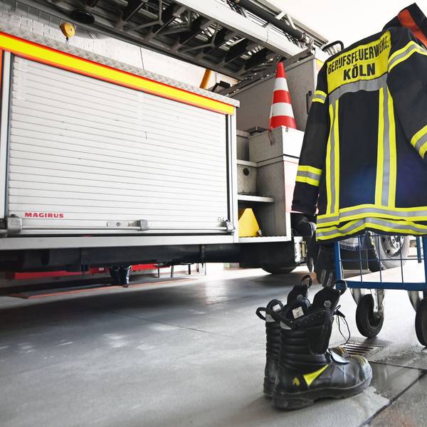 Eine Feuerwehruniform hängt über einem Haken, im Hintergrund steht eine Feuerwehrauto.