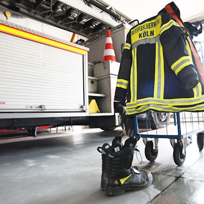 Eine Feuerwehruniform hängt über einem Haken, im Hintergrund steht eine Feuerwehrauto.