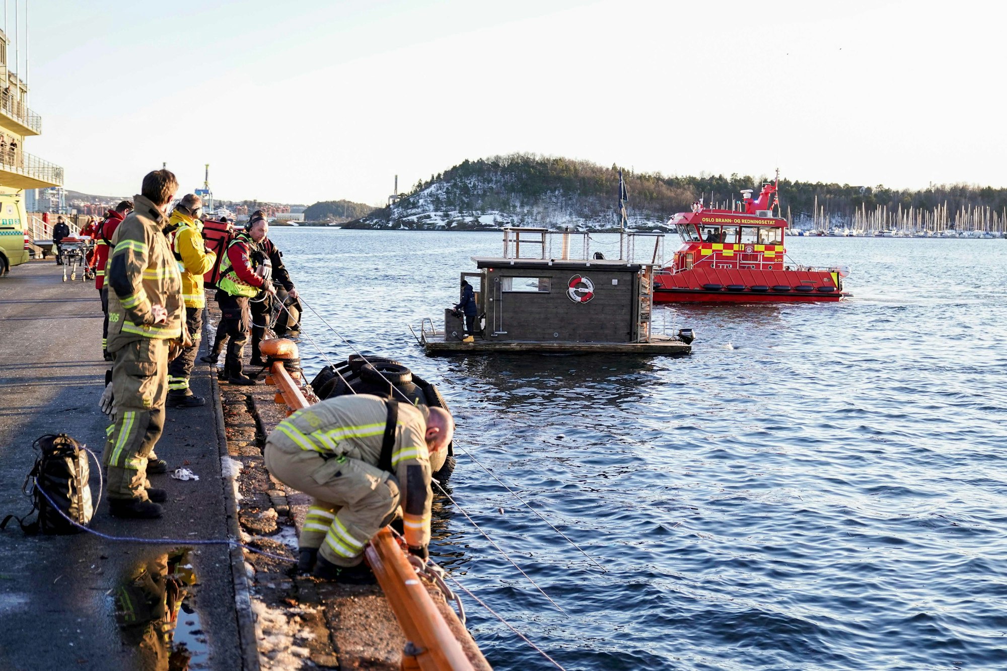 Einsatzkräfte ziehen ein untergegangenes Auto an Land. Auf dem Wasser des Fjords von Oslo treiben eine schwimmende Sauna und ein Rettungsboot.