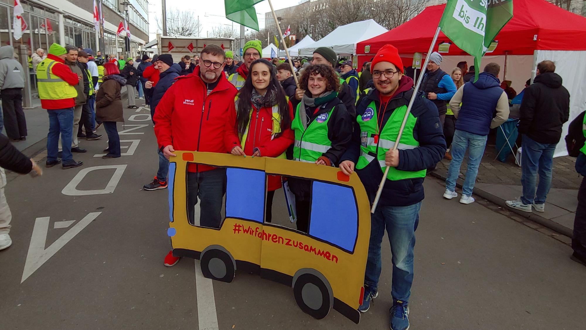 Zu sehen sind Streikende der KVB und der Umweltgruppe „Fridays for Future“ mit einem Plakat aus Pappe, das einen Bus darstellt. Er trägt die Aufschrift „#Wir fahrenzusammen“.