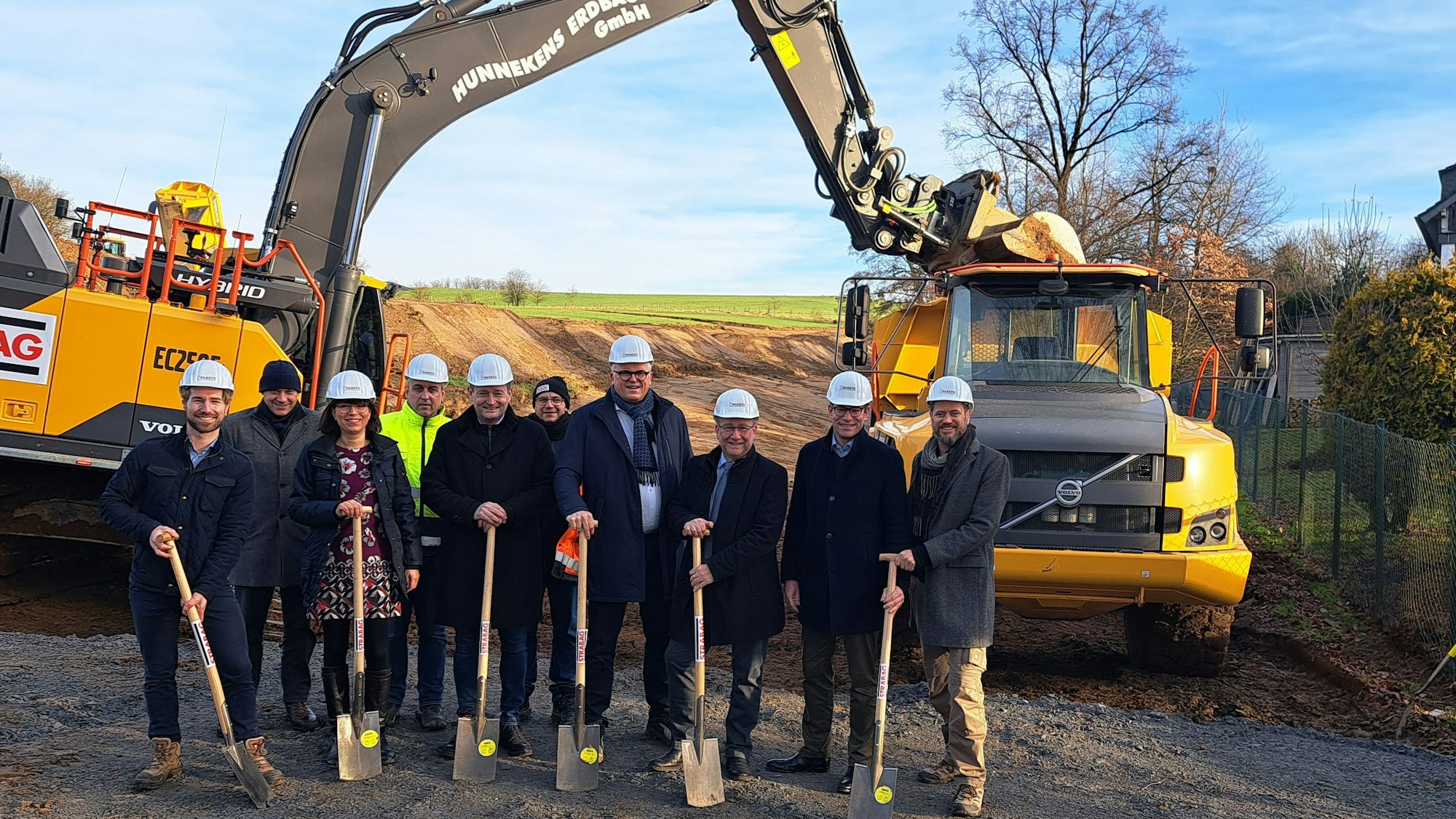 Das Foto zeigt eine Gruppe mit Bauarbeiterhelmen und Spaten, dahinter zwei Baufahrzeuge.
