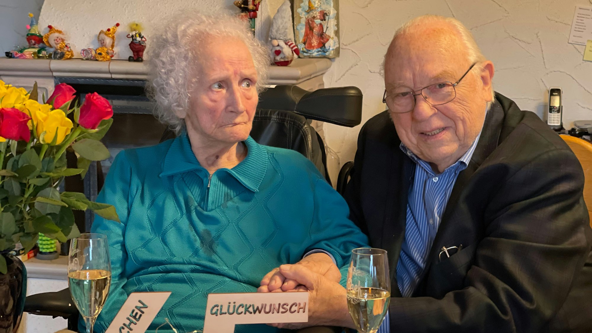 Hildegard und Horst Ziethen sitzen an einem Tisch, auf dem ein Strauß gelber und roter Rosen steht. Vor ihnen stehen Sektgläser.