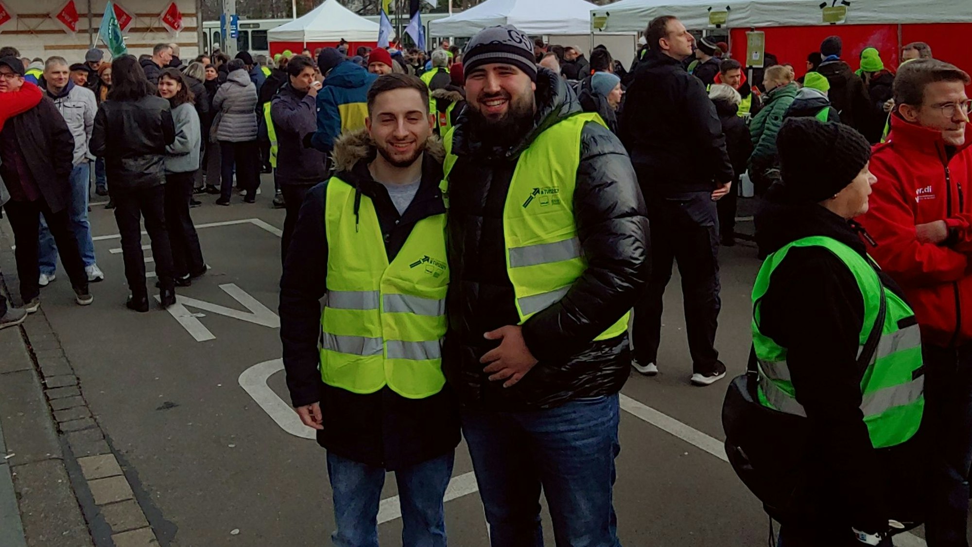 Zwei Streikende der KVB stehen vor weiteren Mitarbeitenden und aufbauten Zelten.