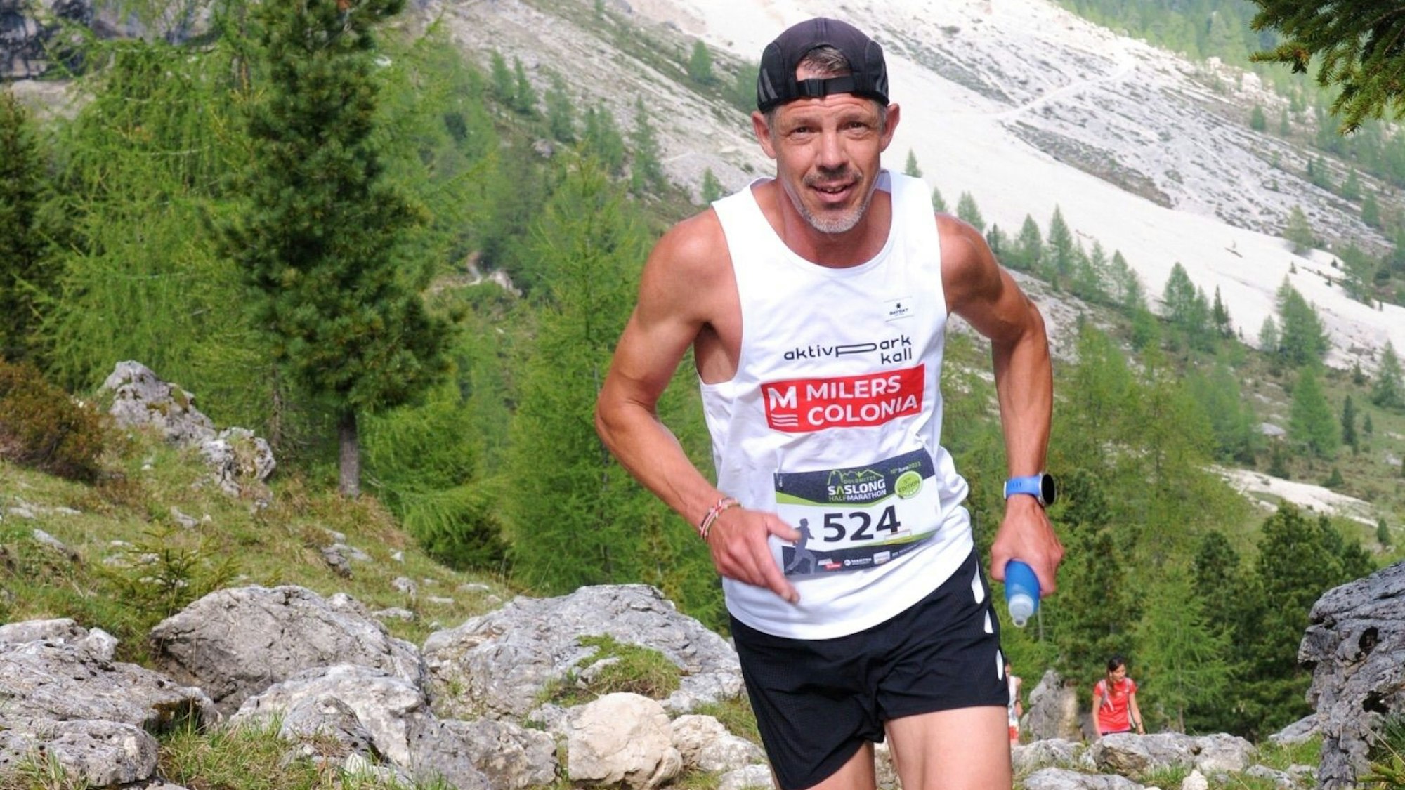 Markus Mey läuft einen Berg hinauf und lächelt dabei. Seine Kappe hat er mit dem Schirm nach hinten aufgesetzt. In der Hand hält er eine Trinkflasche.